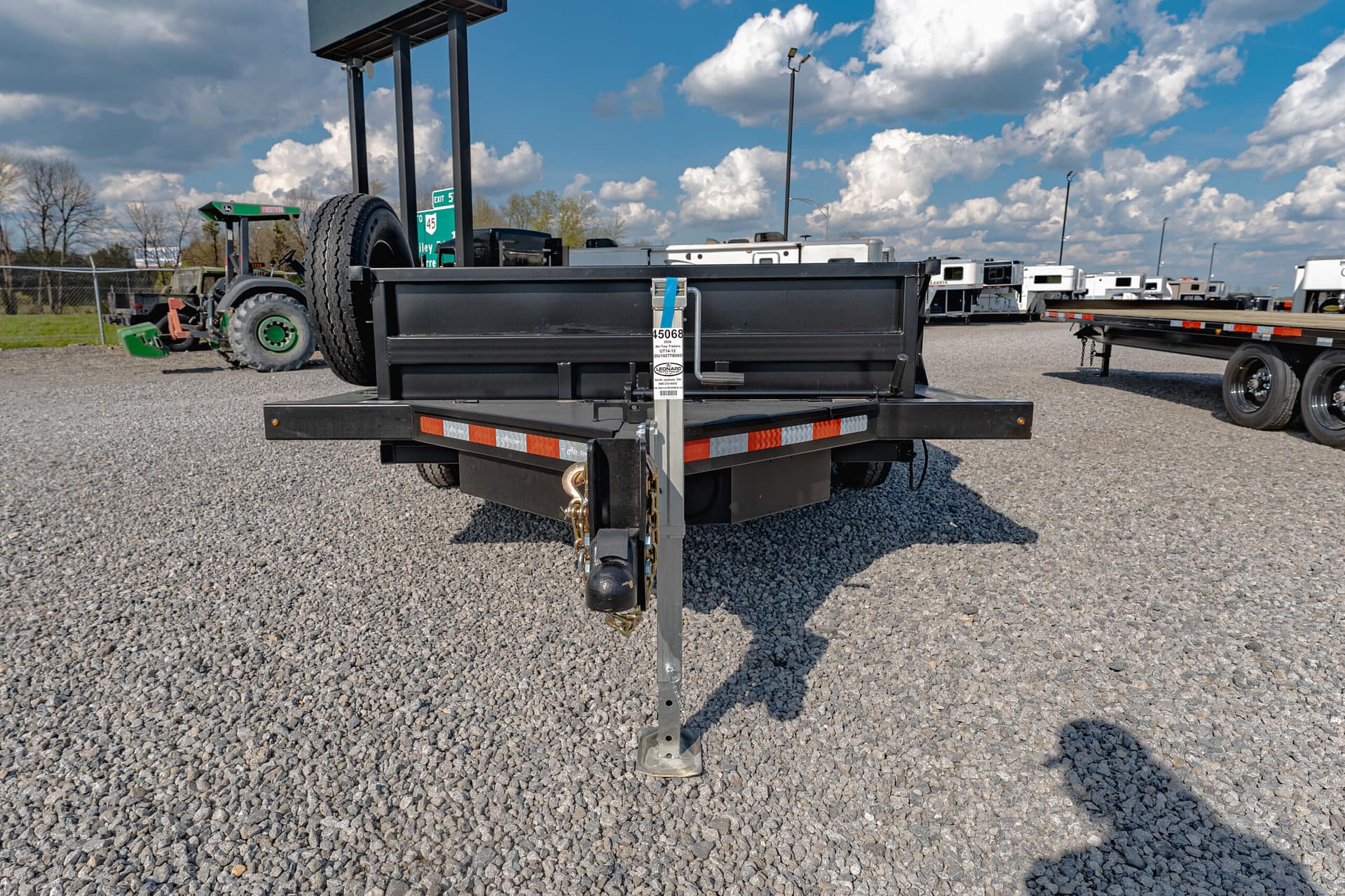 Close-up of a black trailer hitch and support jack on a gravel lot with other trailers in the background under a blue sky.