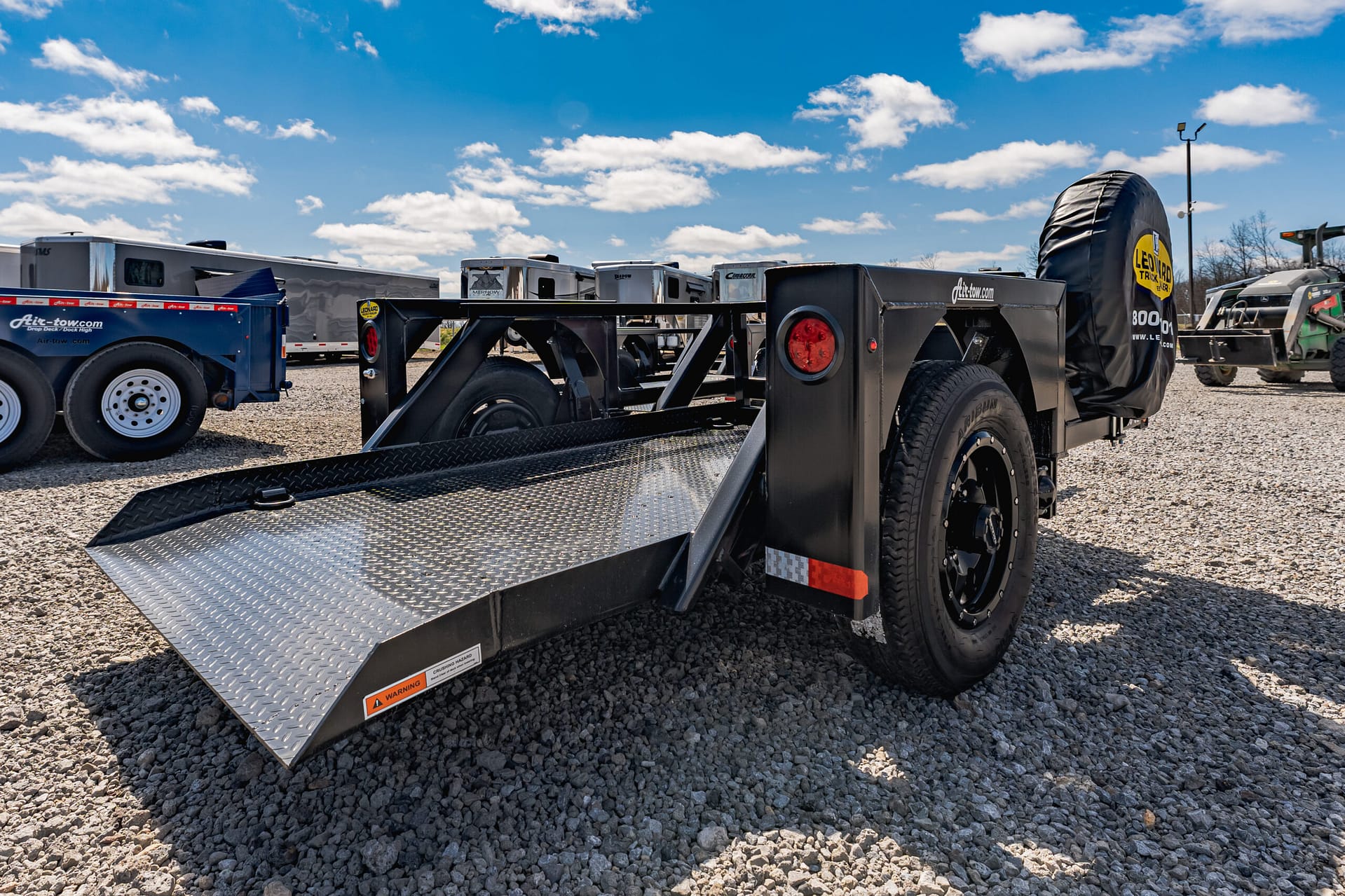 Close-up of a black steel trailer with a diamond-plate loading ramp on a gravel lot, with other trailers and a bright blue sky in the background, conveying a vehicle-hauling yard.