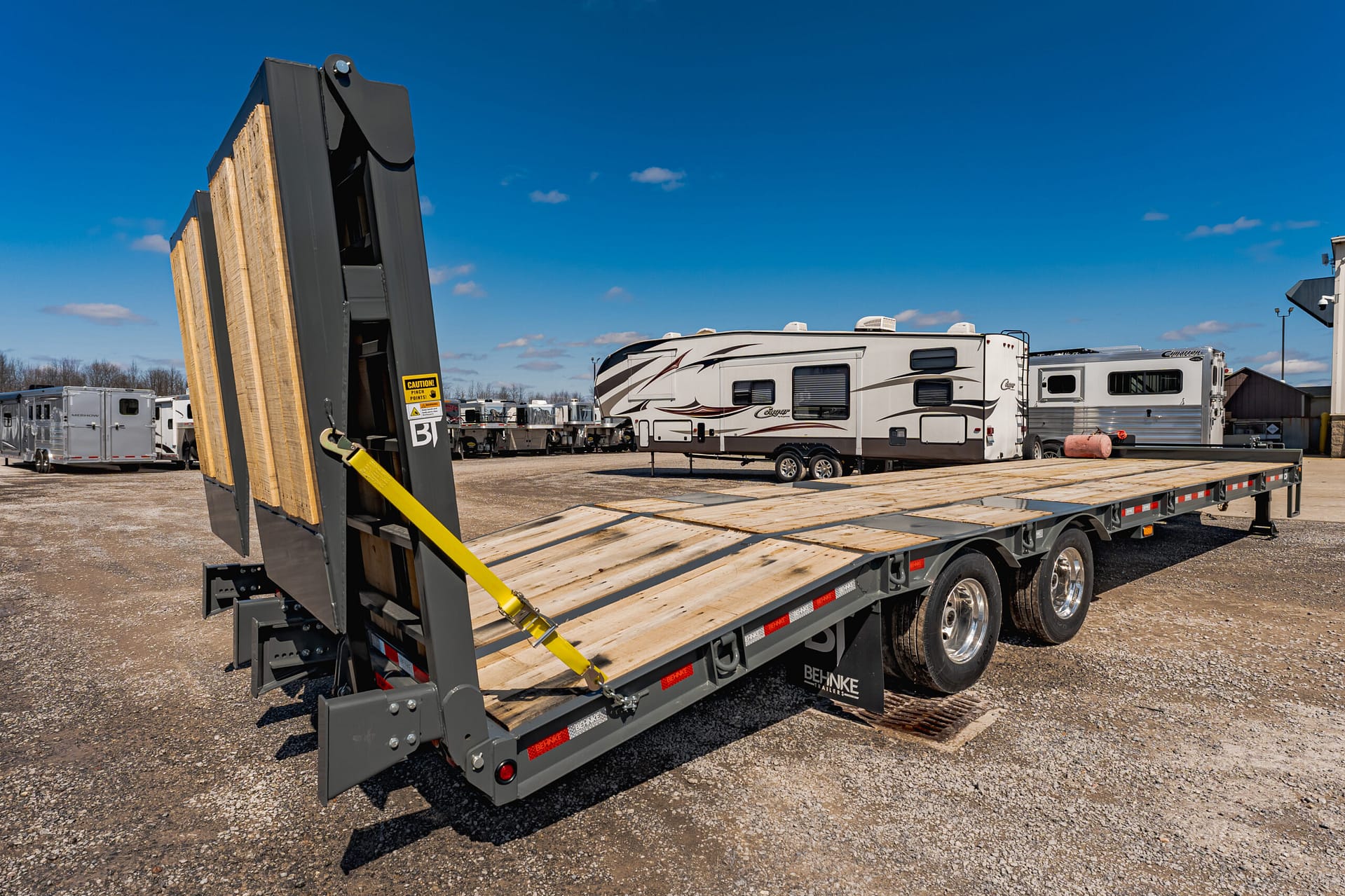Flatbed trailer with a wooden deck and a raised ramp in an RV storage yard under a clear blue sky.