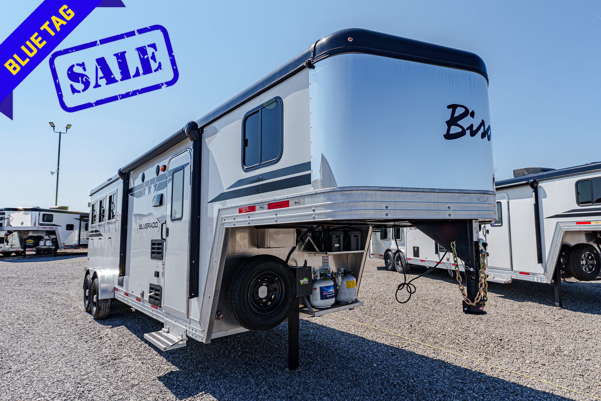 RV trailer on a gravel lot with a large blue 'SALE' stamp and a diagonal 'BLUE TAG' banner in the top-left corner
