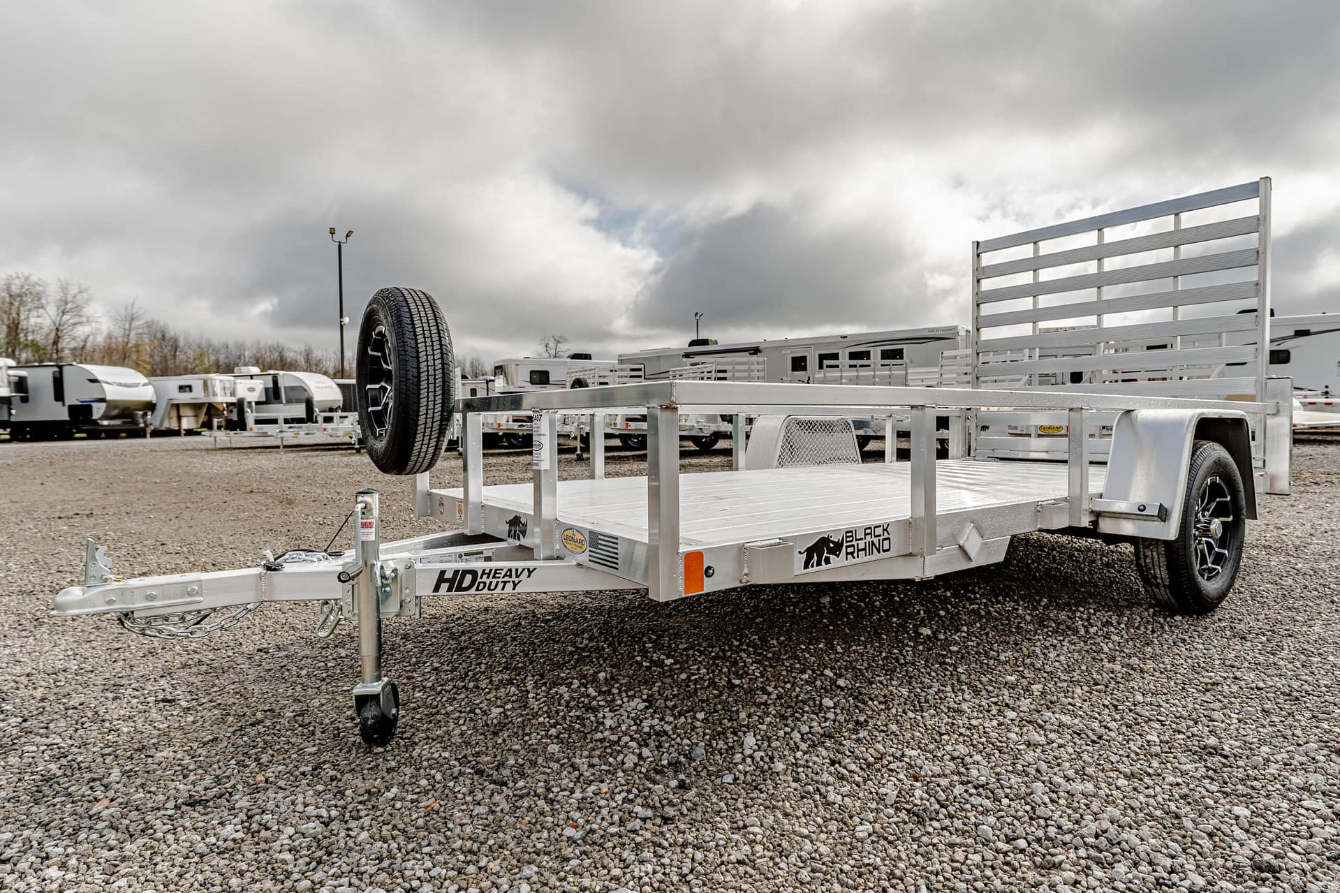 Aluminum flatbed utility trailer with a spare tire mounted on the front left, parked on a gravel lot among other trailers in a storage yard.