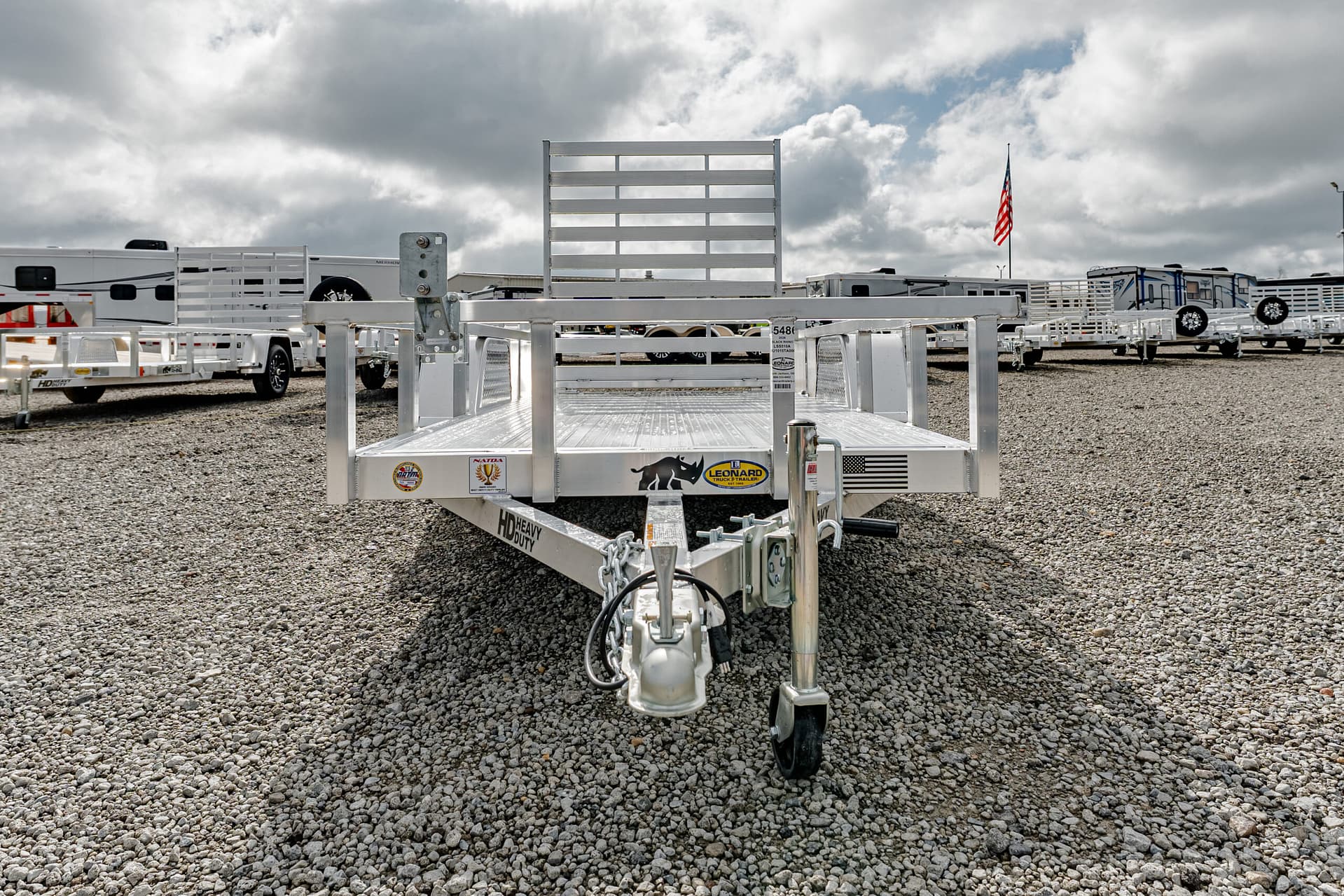 Front view of a metal utility trailer on a gravel lot, with an American flag and other trailers in the background under a cloudy sky.