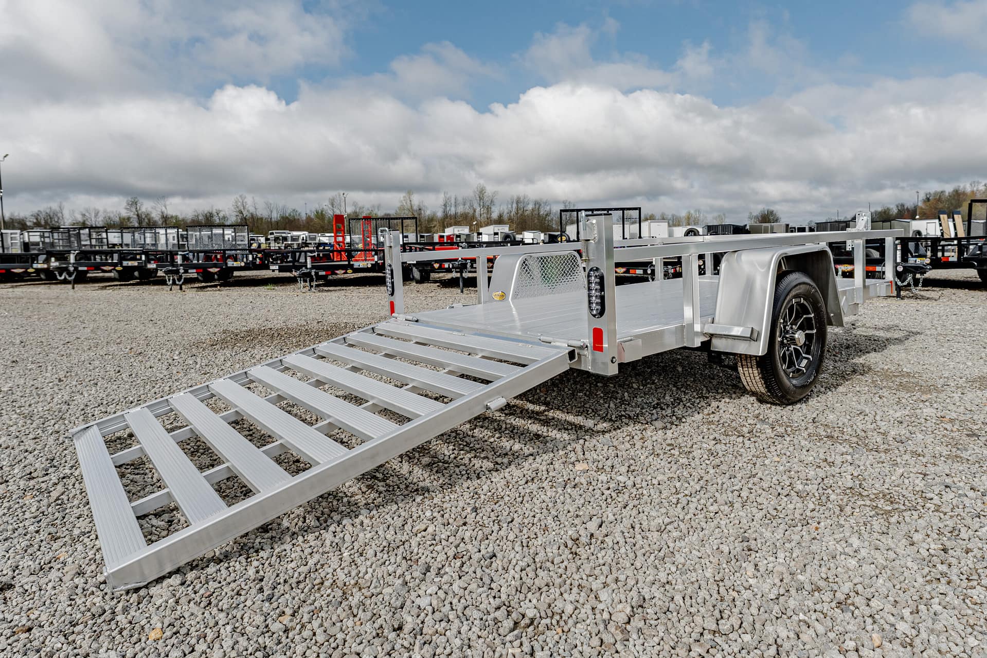 Aluminum trailer with ramp extended on a gravel lot, several other trailers in the background under a cloudy sky