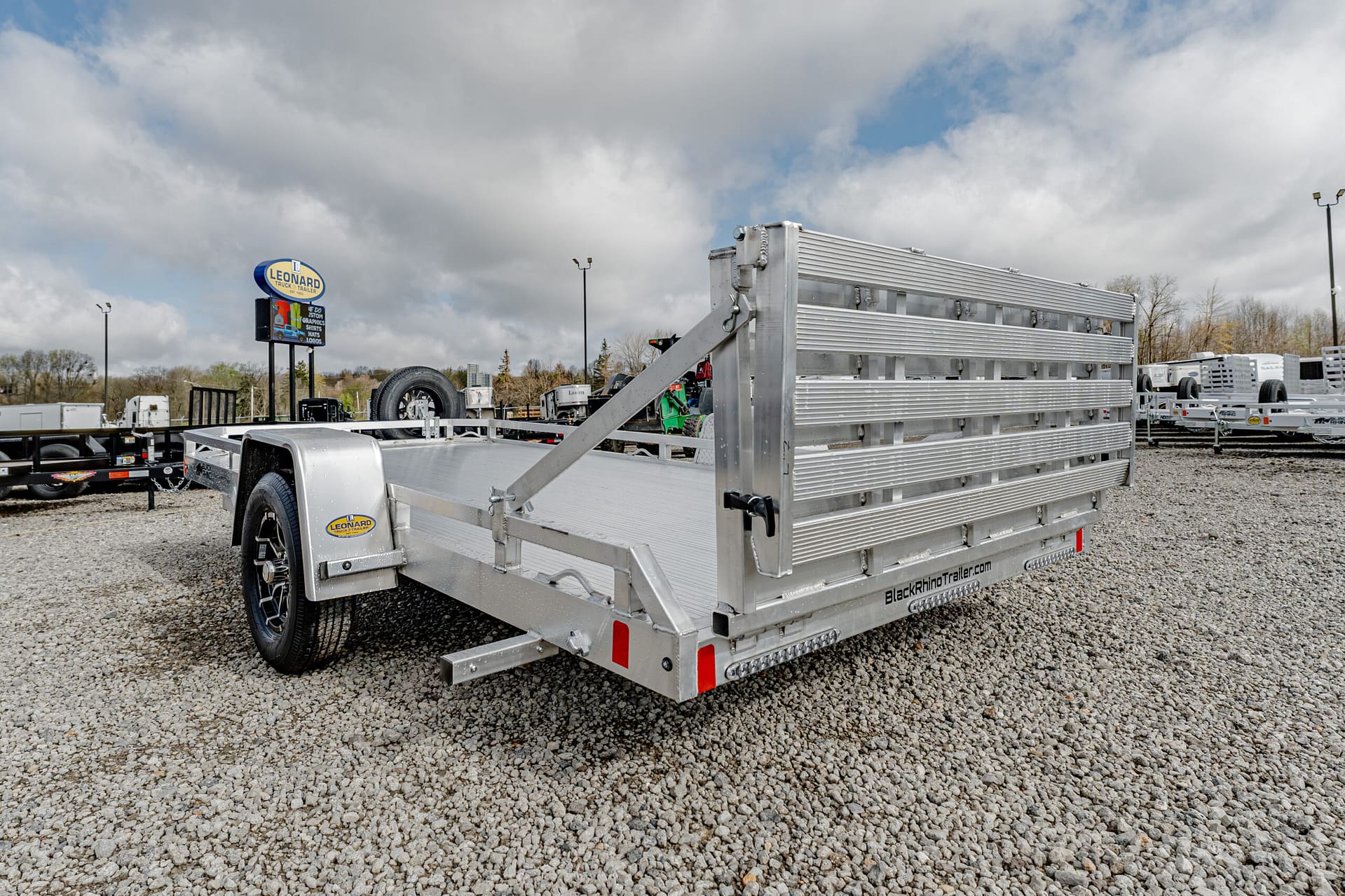 Aluminum flatbed trailer with a fold-down ramp in a gravel lot, brand visible on side (Black Rhino Trailer) and a spare tire at the front left.