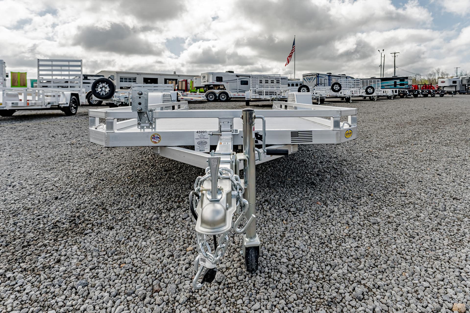 Close-up of a metal trailer hitch and chains in a gravel lot, with other trailers and RVs in the background under a cloudy sky.