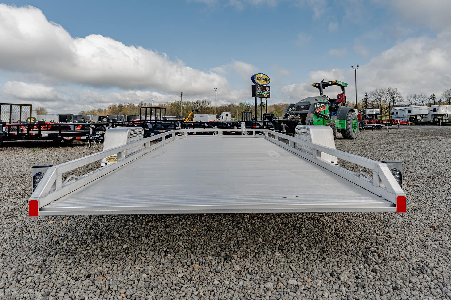 Front-facing view of a silver aluminum flatbed trailer in a gravel truck yard with other trailers and equipment in the background under a blue sky.
