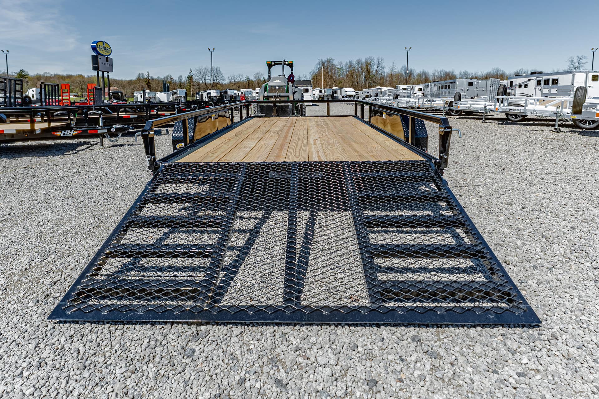 Front view of a black metal flatbed trailer with a wooden deck in a gravel lot, surrounded by other trailers.]','Wide shot of a trailer lot with numerous white and silver trailers and equipment in the background.