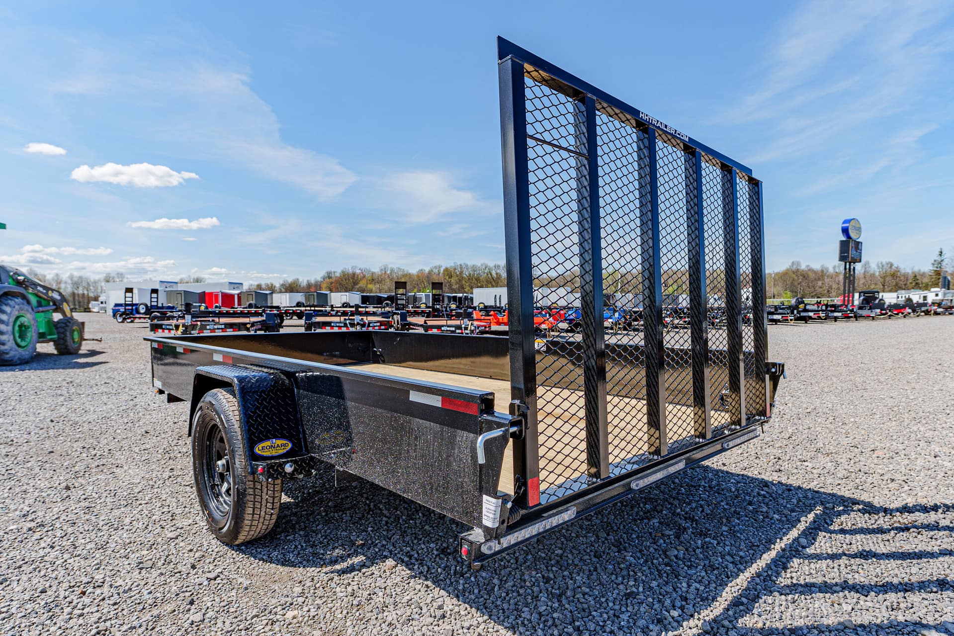 Close-up of a black flatbed trailer with a tall metal mesh gate, parked on a gravel lot with more trailers in the background under a blue sky