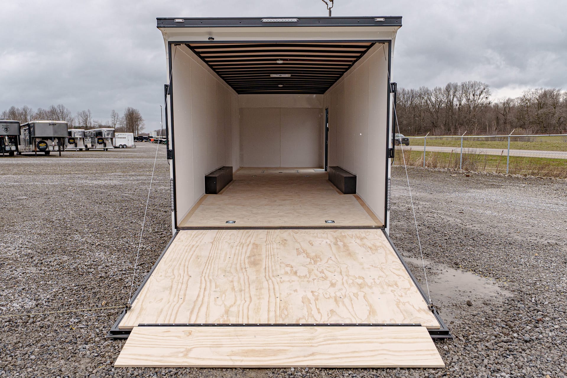 Open empty cargo trailer with ramp down, plywood floor, and bare interior in a gravel lot.
