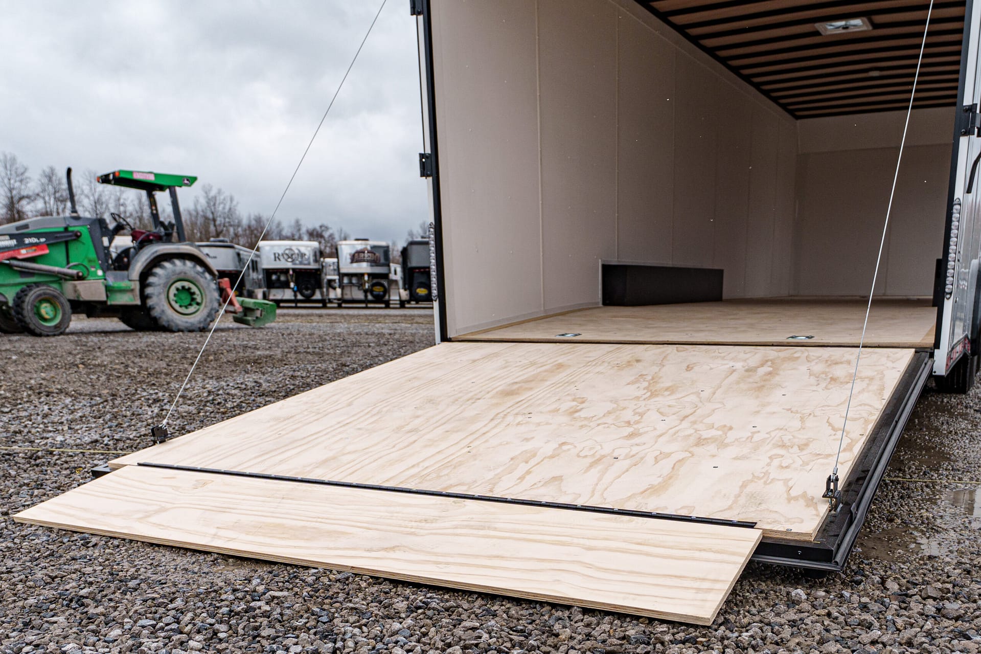 Open trailer doorway with a wooden ramp, empty interior, and a gravel lot with machinery in the background on a cloudy day, including a green tractor.