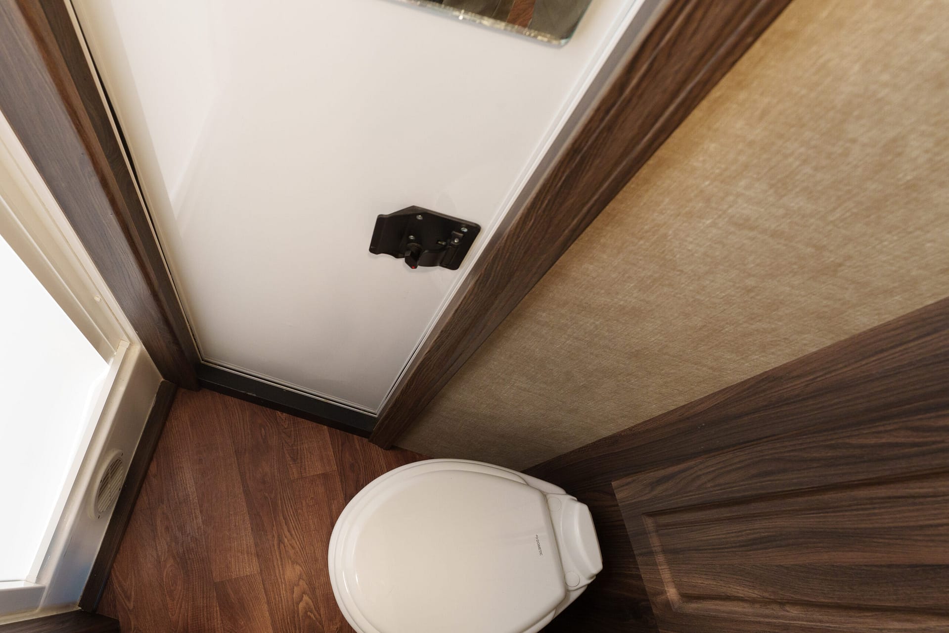 Small bathroom area in a camper or RV, featuring a white door with a black latch, wood trim, and a closed toilet seat in the foreground.