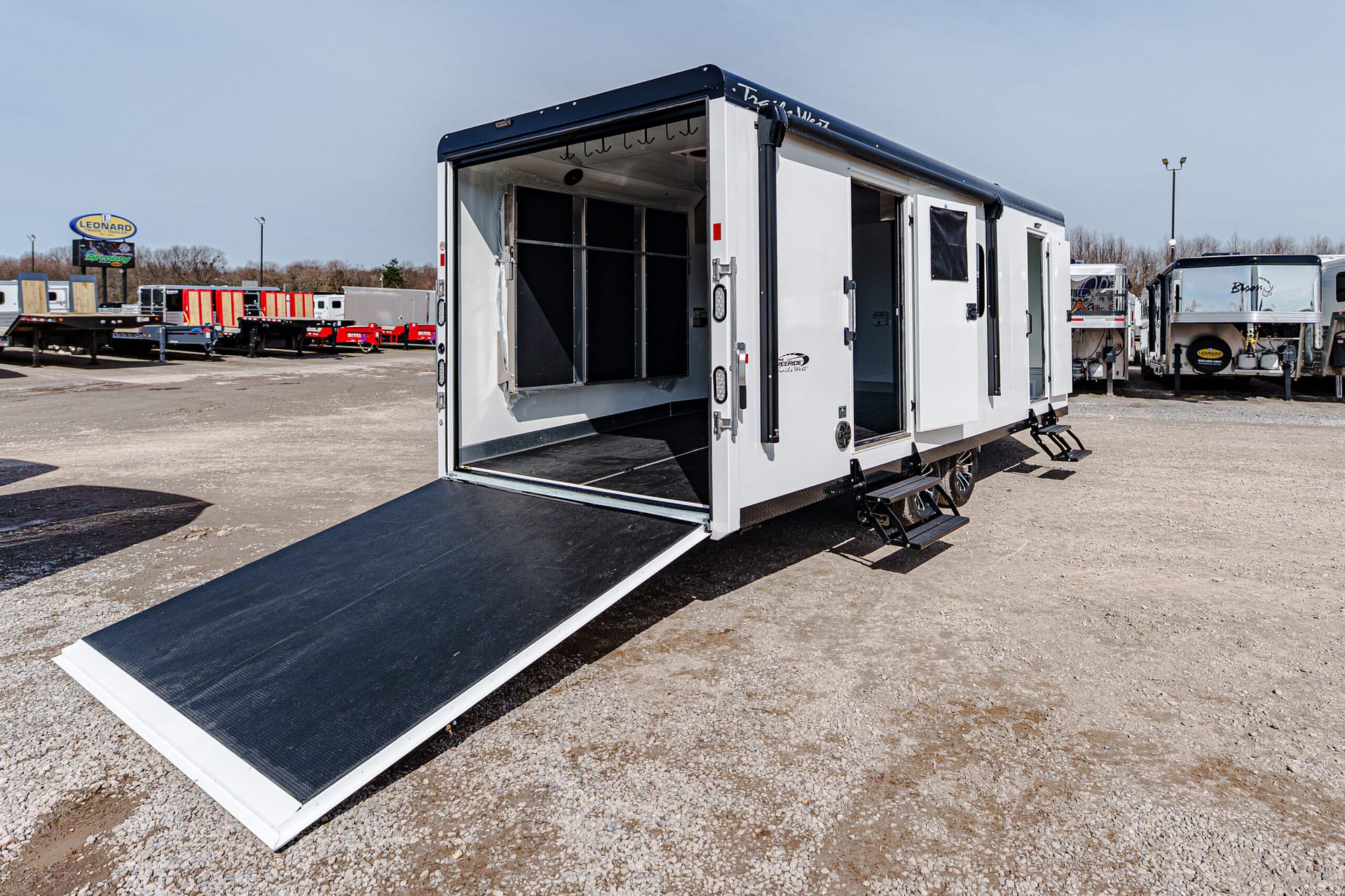 White utility trailer with an extended black ramp on a gravel lot, other trailers in the background