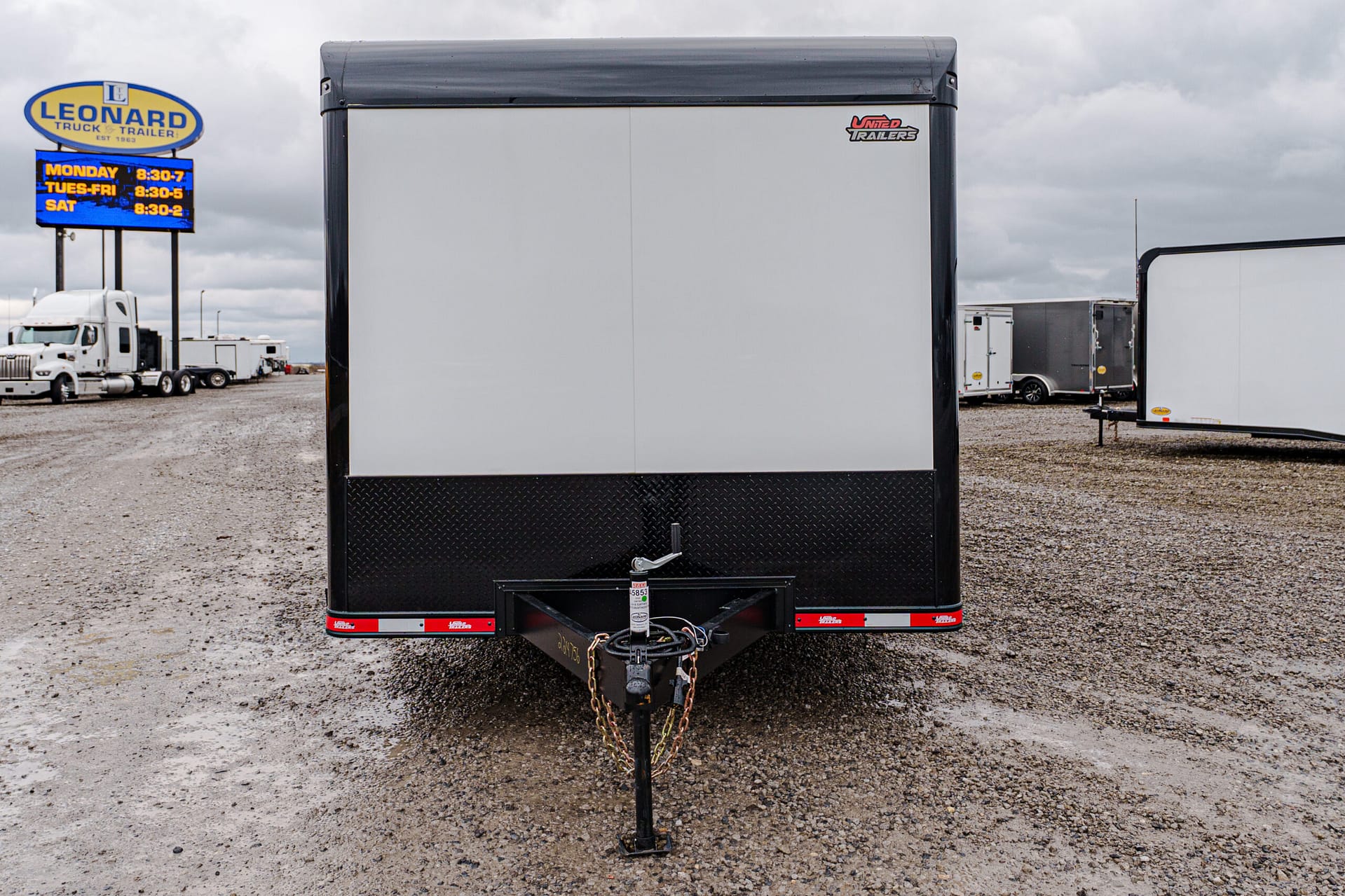 Rear view of a large silver trailer in a gravel truck yard, with a hitch and chains visible and other trailers in the background.