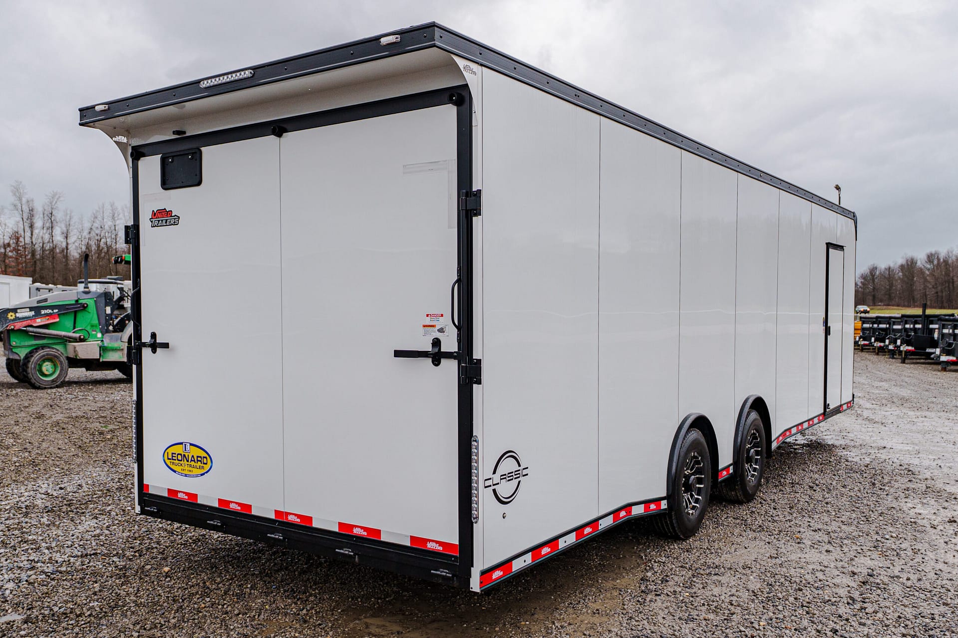 White enclosed cargo trailer with black framing and double back doors on a gravel lot; visible brand stickers on the door and side edges.