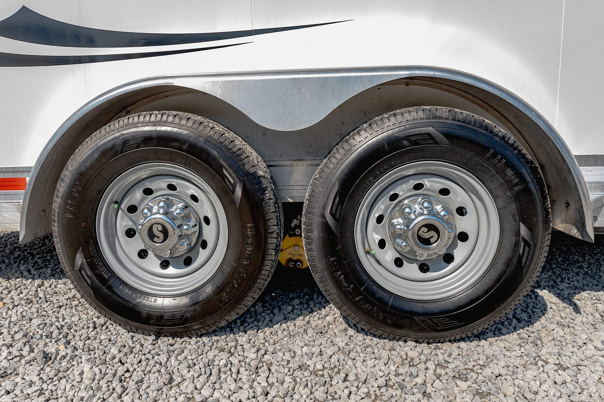 Close-up of two trailer wheels with chrome hubs under a curved silver fender on a gravel surface