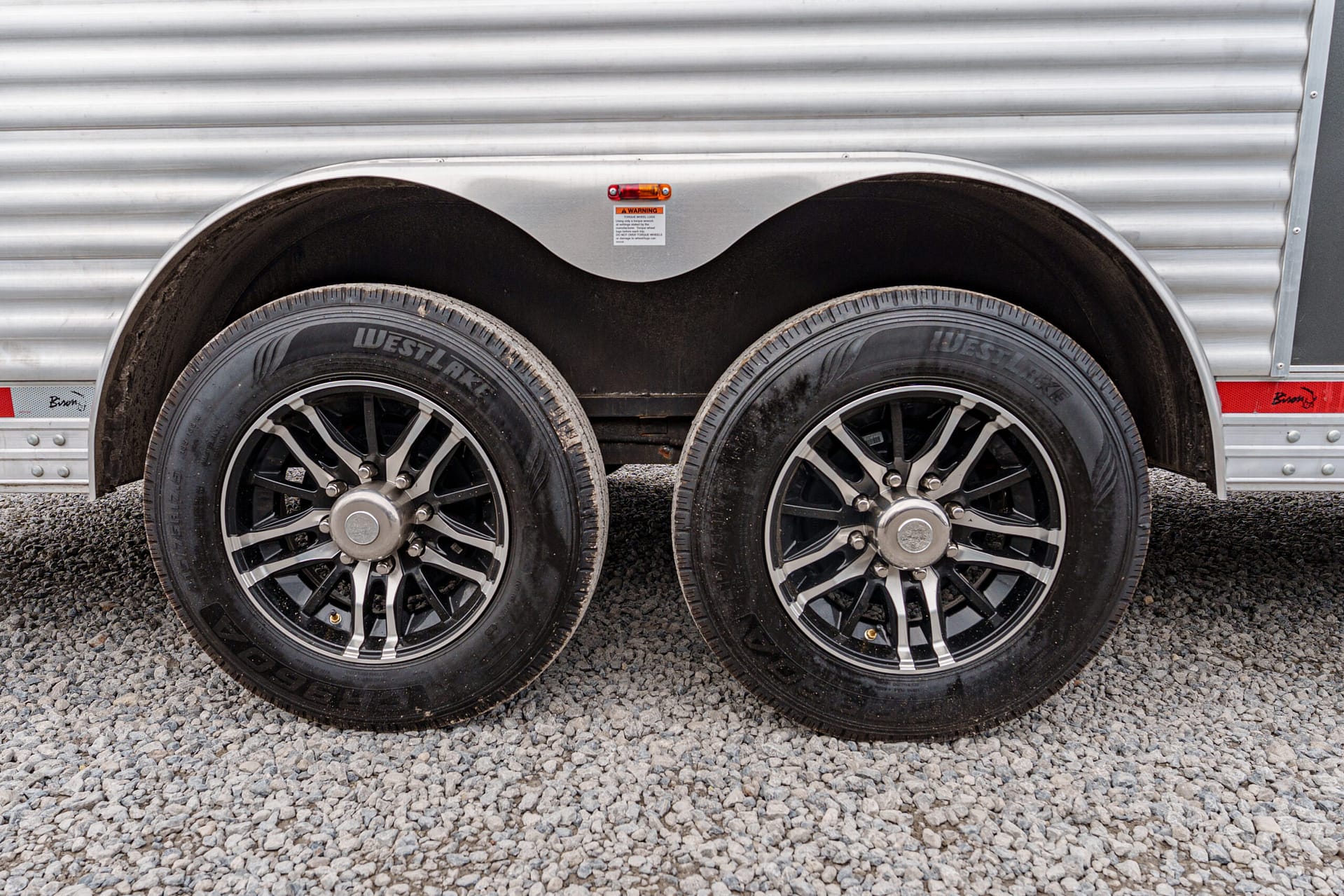 Dual trailer wheels with black alloy rims under a silver corrugated trailer body on gravel surface