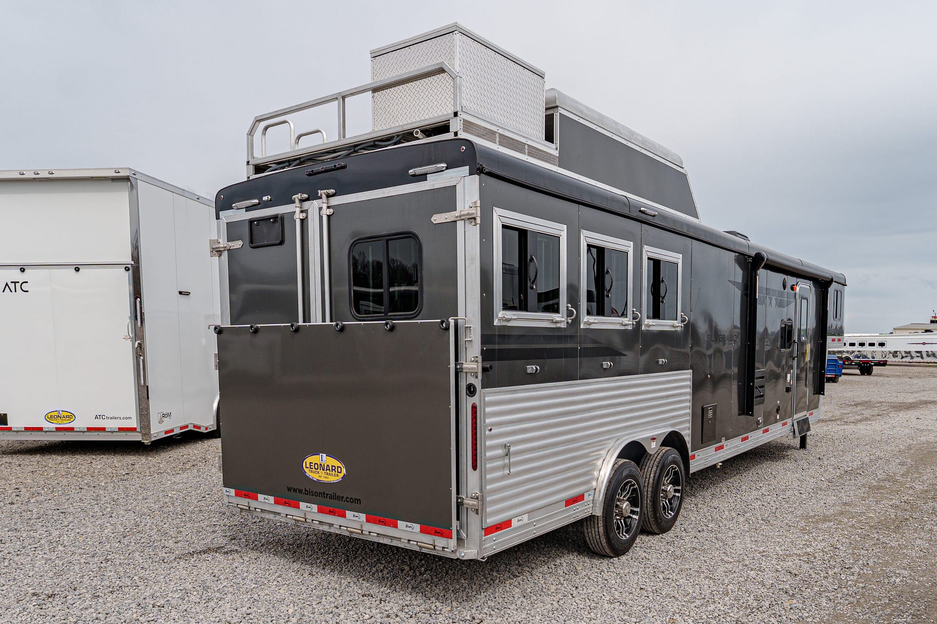 Long dark trailer with aluminum trim, multiple side windows, and a diamond-plate storage box on top, parked on gravel with other trailers in the background.