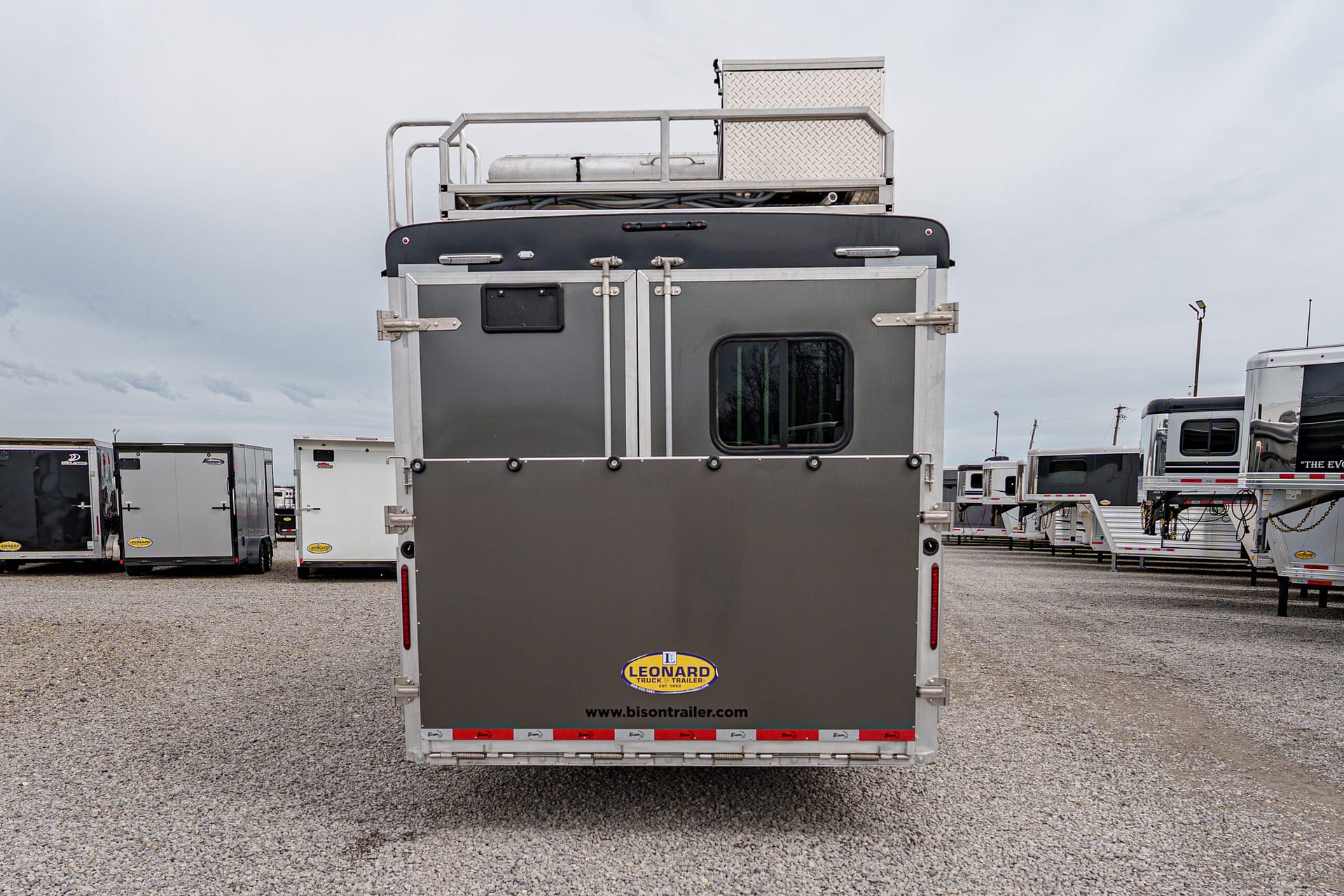 Rear view of a gray utility trailer with metal framing, a diamond-plate top box, and a Leonard trailer logo on the back door edge, in a lot of trailers