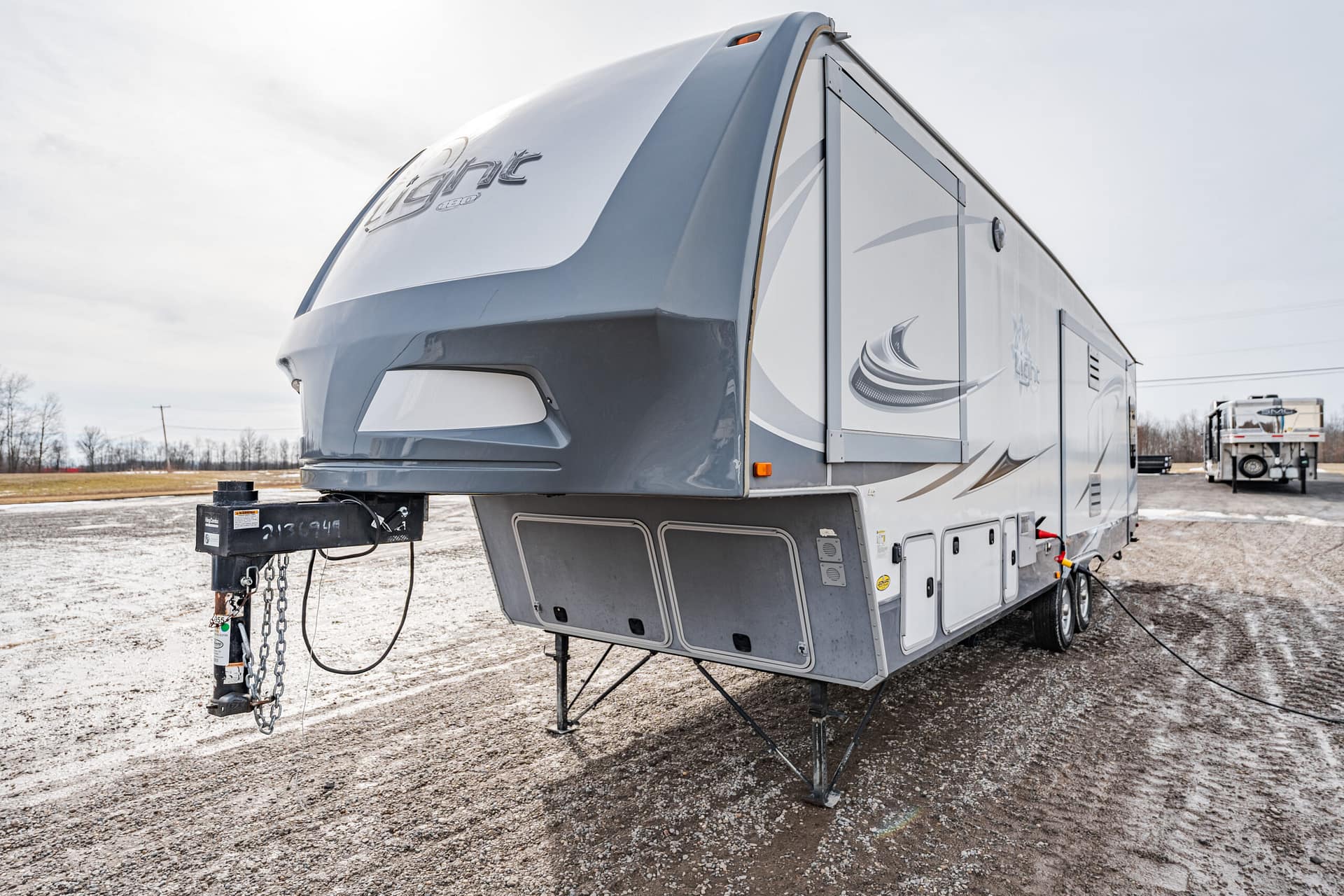 A large gray and white fifth-wheel RV trailer parked on a gravel lot, showing the hitch and front stabilizers.