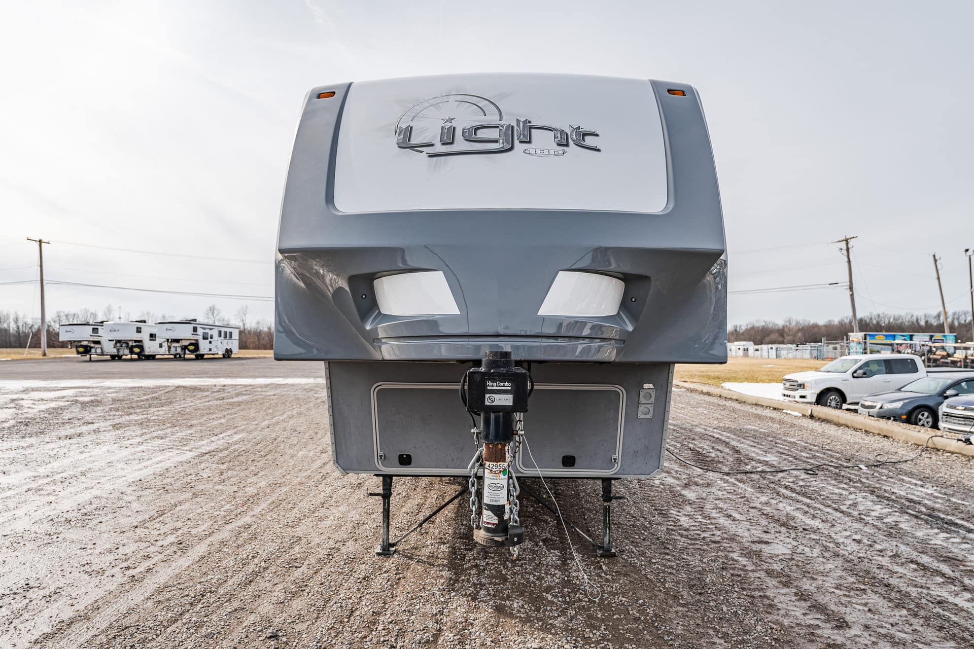 Front view of a gray fifth-wheel trailer with its hitch and stabilizing legs on a gravel lot, logo on the front cap visible