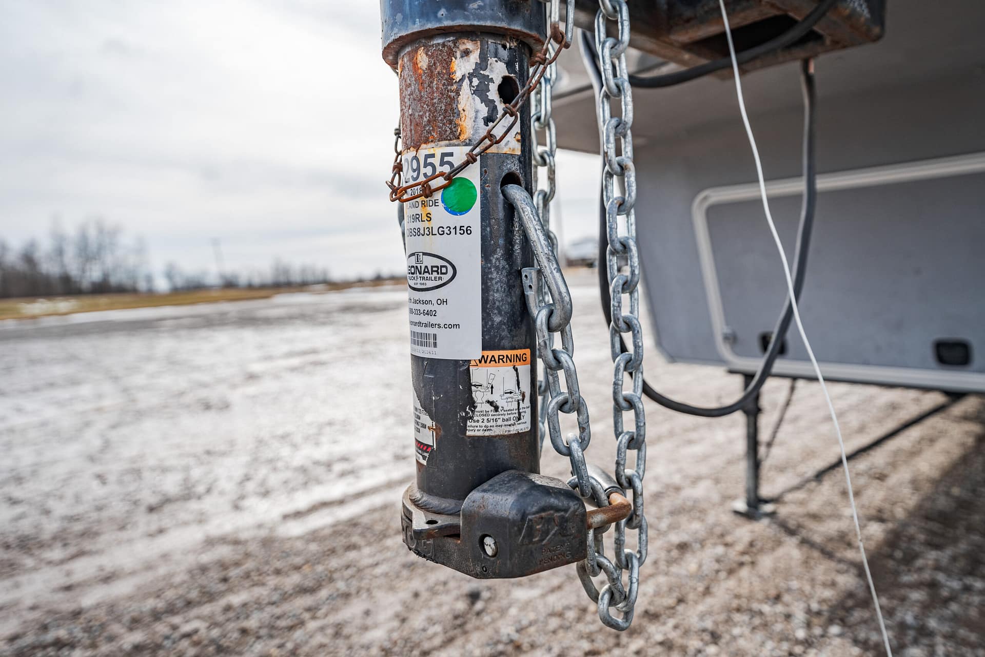 Close-up of a rusty trailer hitch post with chains, a padlock, and warning labels on a gravel lot. Outdoor scene with a trailer tongue in view.