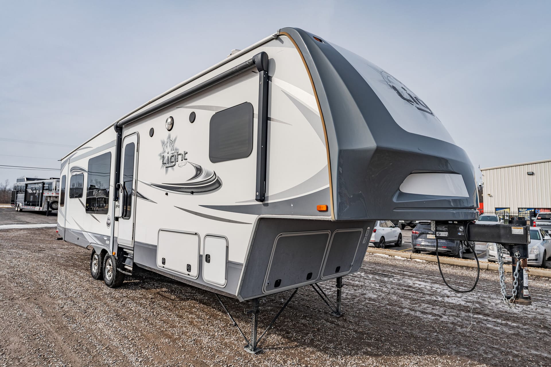 White and gray fifth-wheel RV on a gravel lot with a raised front cap and multiple storage bays visible