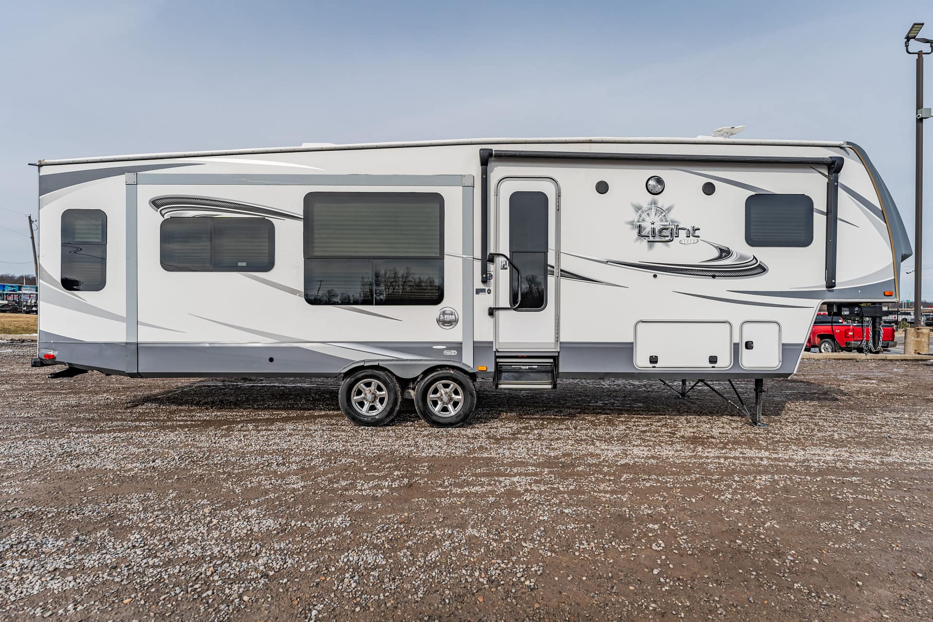 White and gray fifth-wheel travel trailer parked on gravel, with multiple windows and an entry door on the side.
