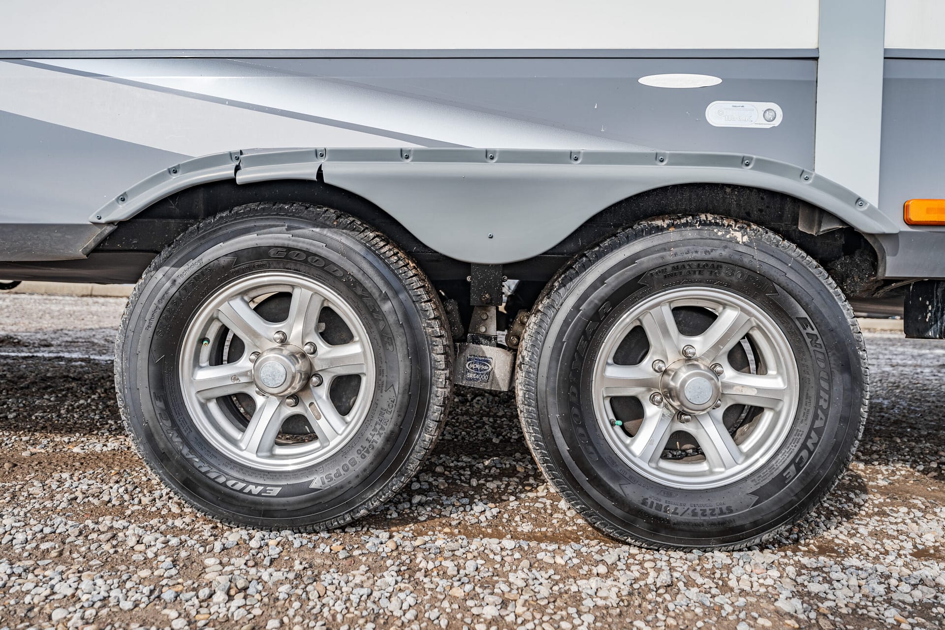 Dual-axle trailer wheels with silver rims on a gravel surface under a gray trailer edge.