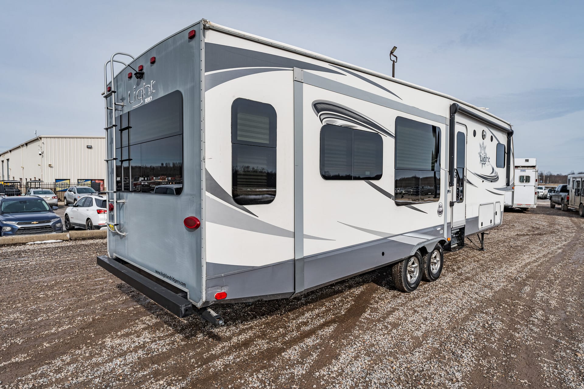 Rear and side view of a large white and gray travel trailer with curved graphics, parked on a gravel lot with other vehicles nearby.