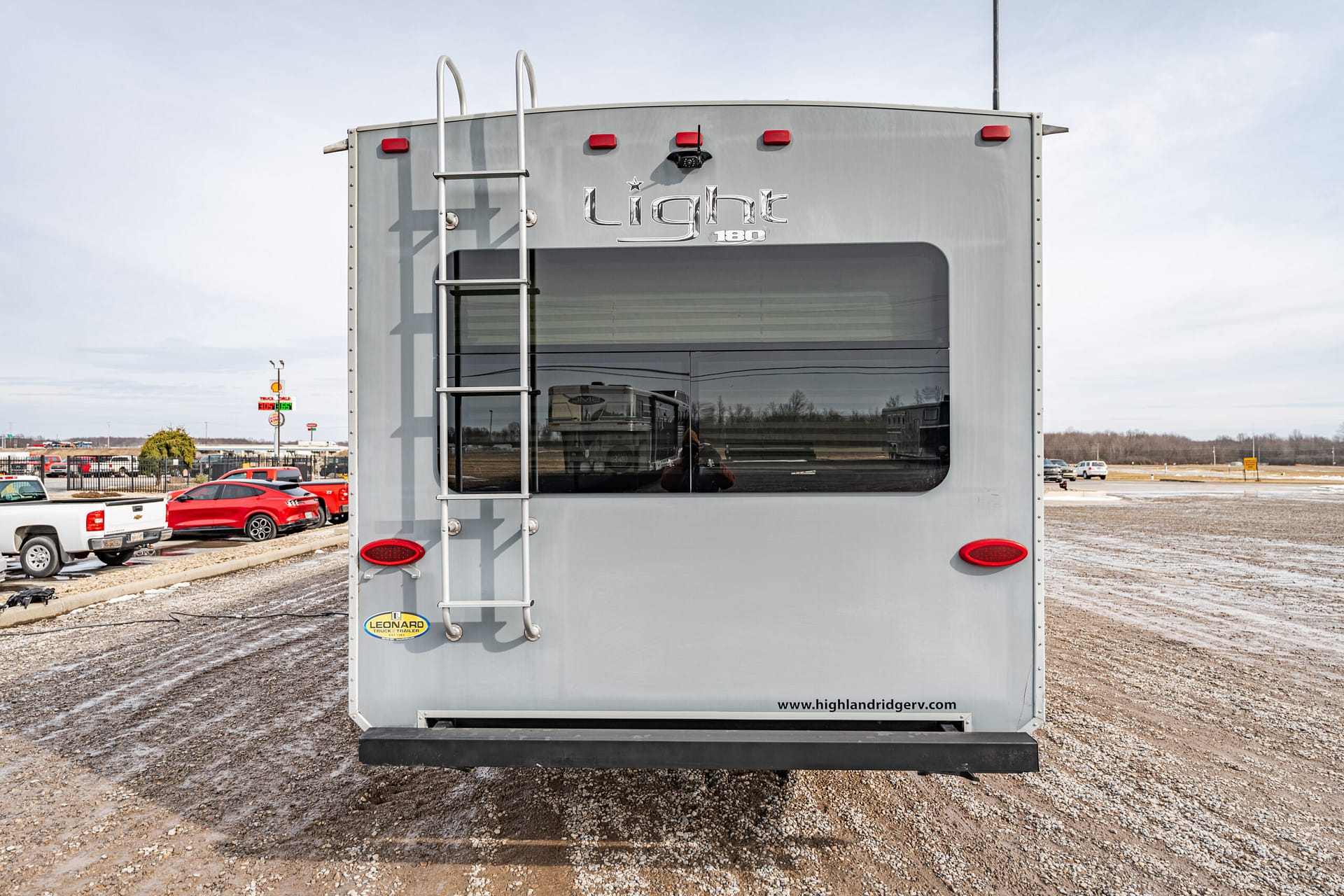 Rear view of a silver Light 180 travel trailer with a vertical ladder and large rear window in a gravel lot. Red taillights visible beneath the window.