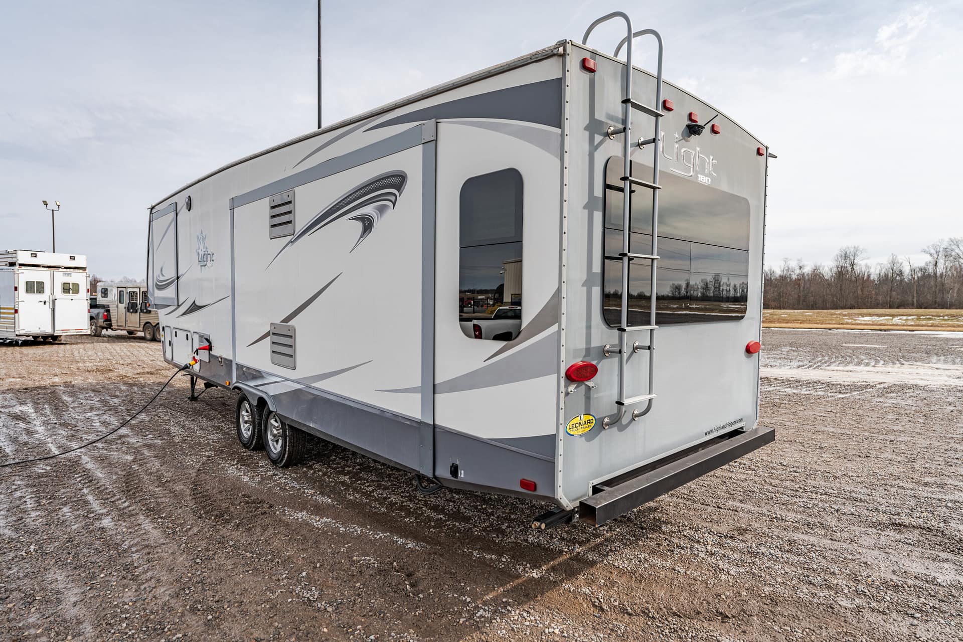 Back of a grey and white travel trailer with a ladder and rear bumper in a gravel lot, other RVs visible in the background