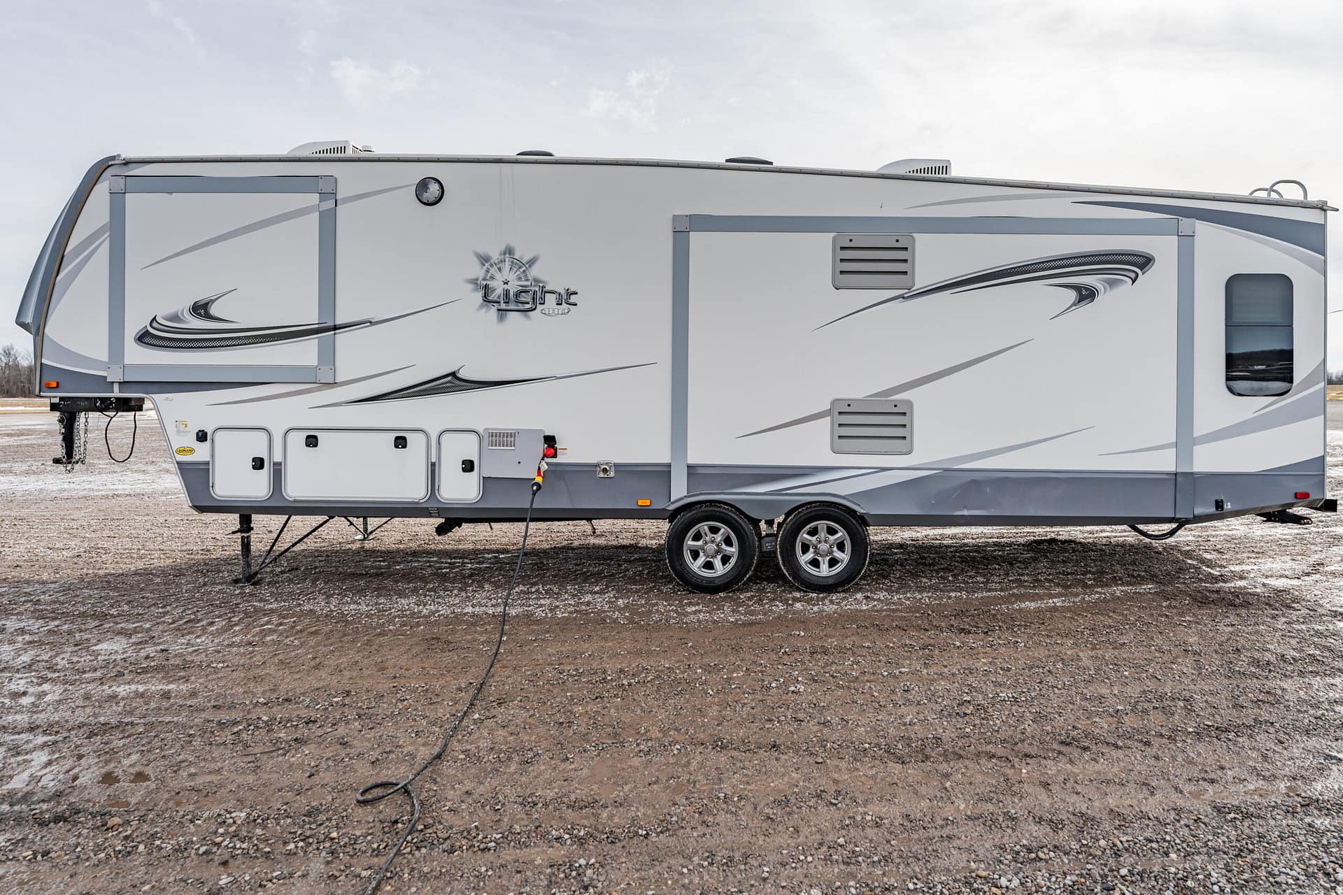 Grey and white travel trailer with graphic swooshes, parked on a gravel lot; power cord plugged into a pedestal nearby.