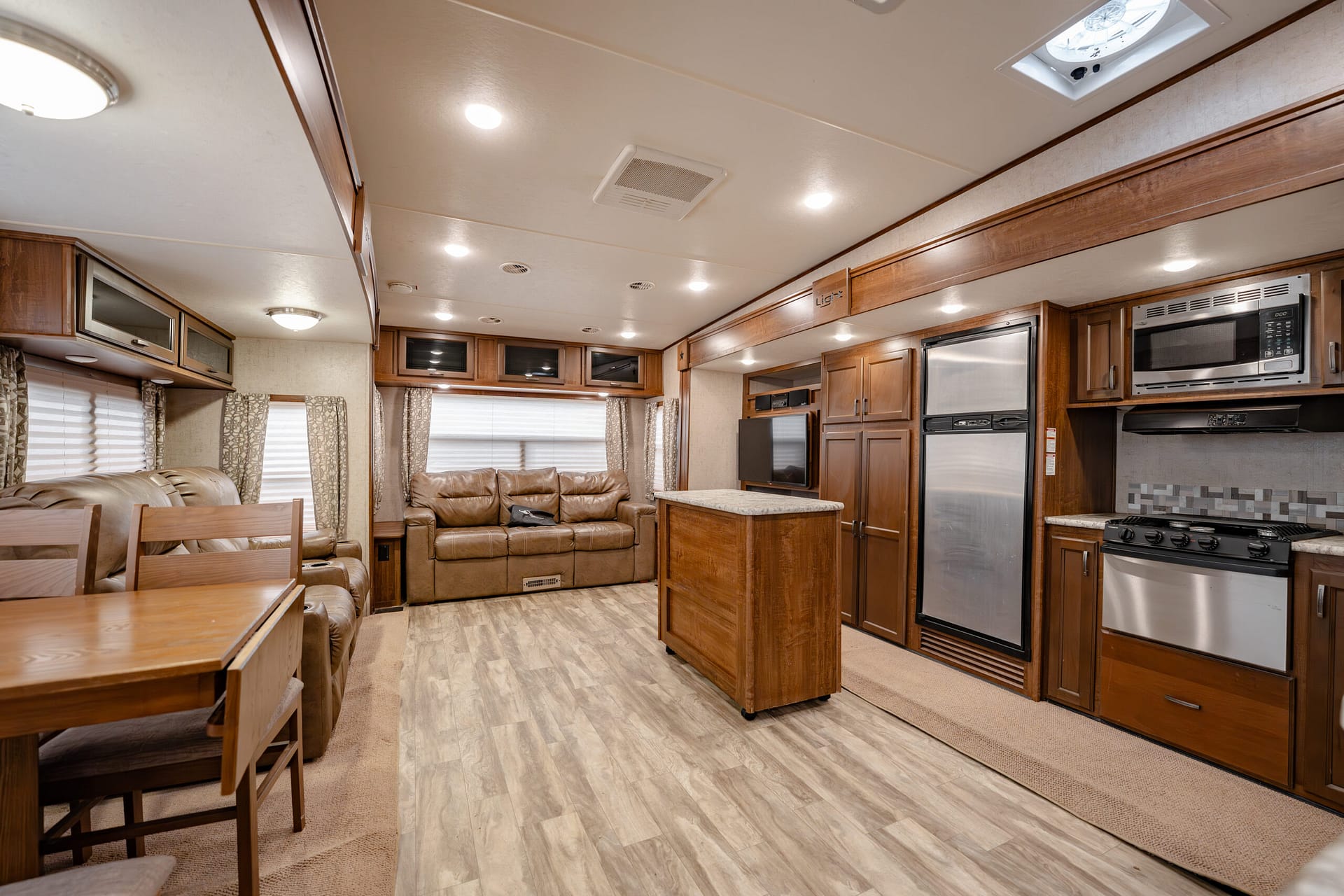 Interior of a modern RV: open living/kitchen area with sofa, dining table, and a central island amid wooden cabinetry and stainless appliances.