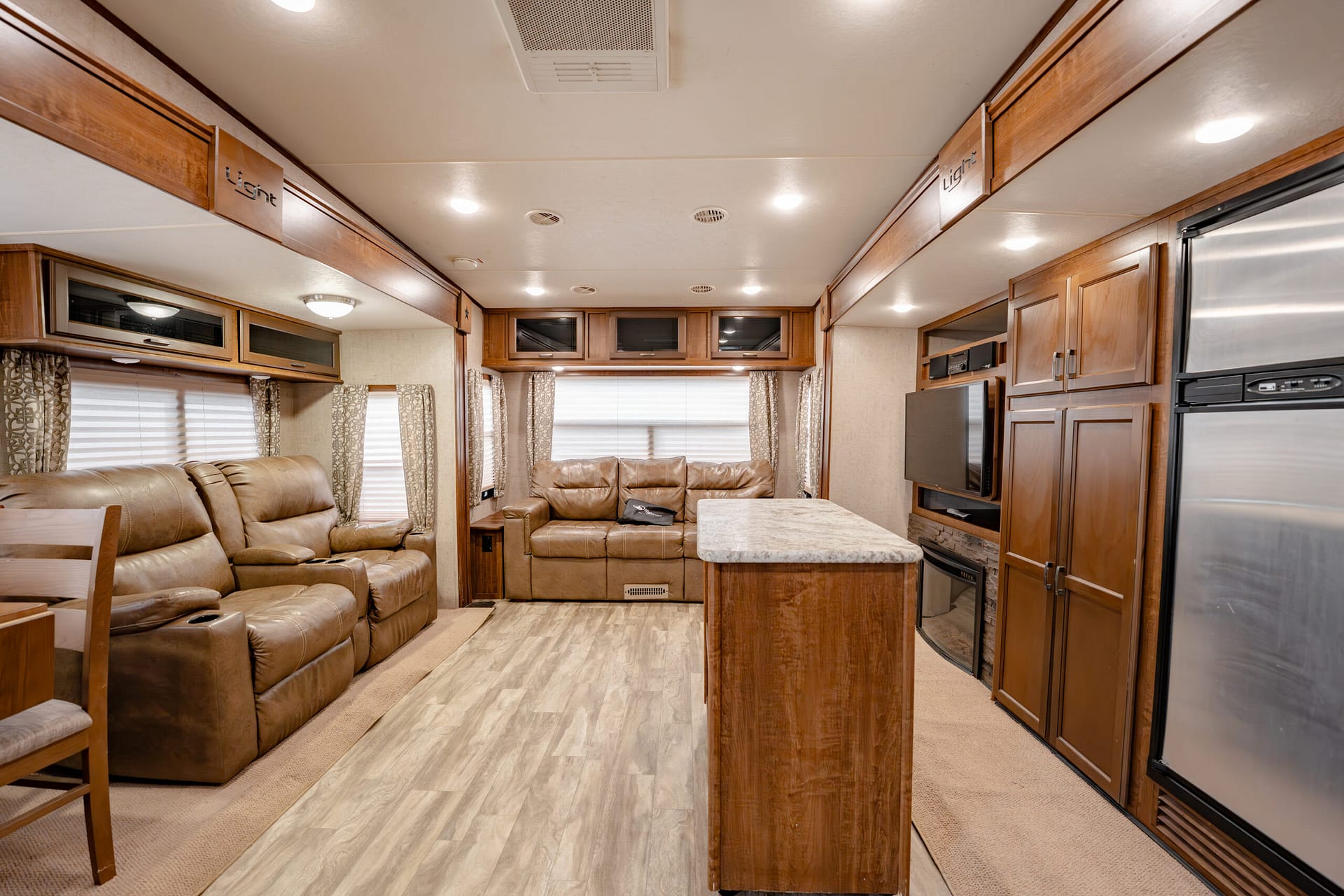 Interior of a spacious RV living area with brown leather sofas, a marble-topped island, and wooden cabinets nearby.