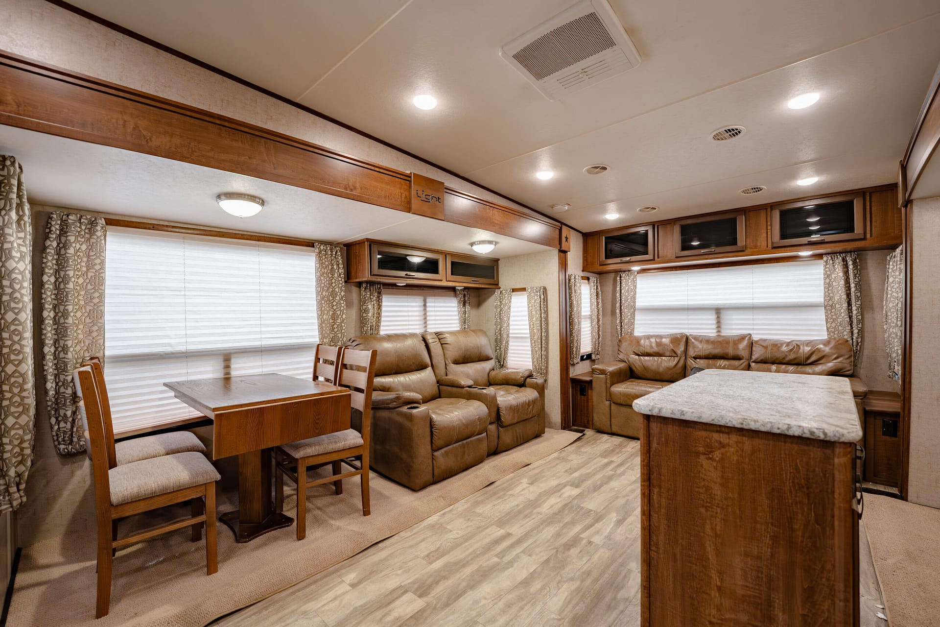RV living space with brown leather recliners, a wooden dining table for four, and a marble-topped island in the foreground.