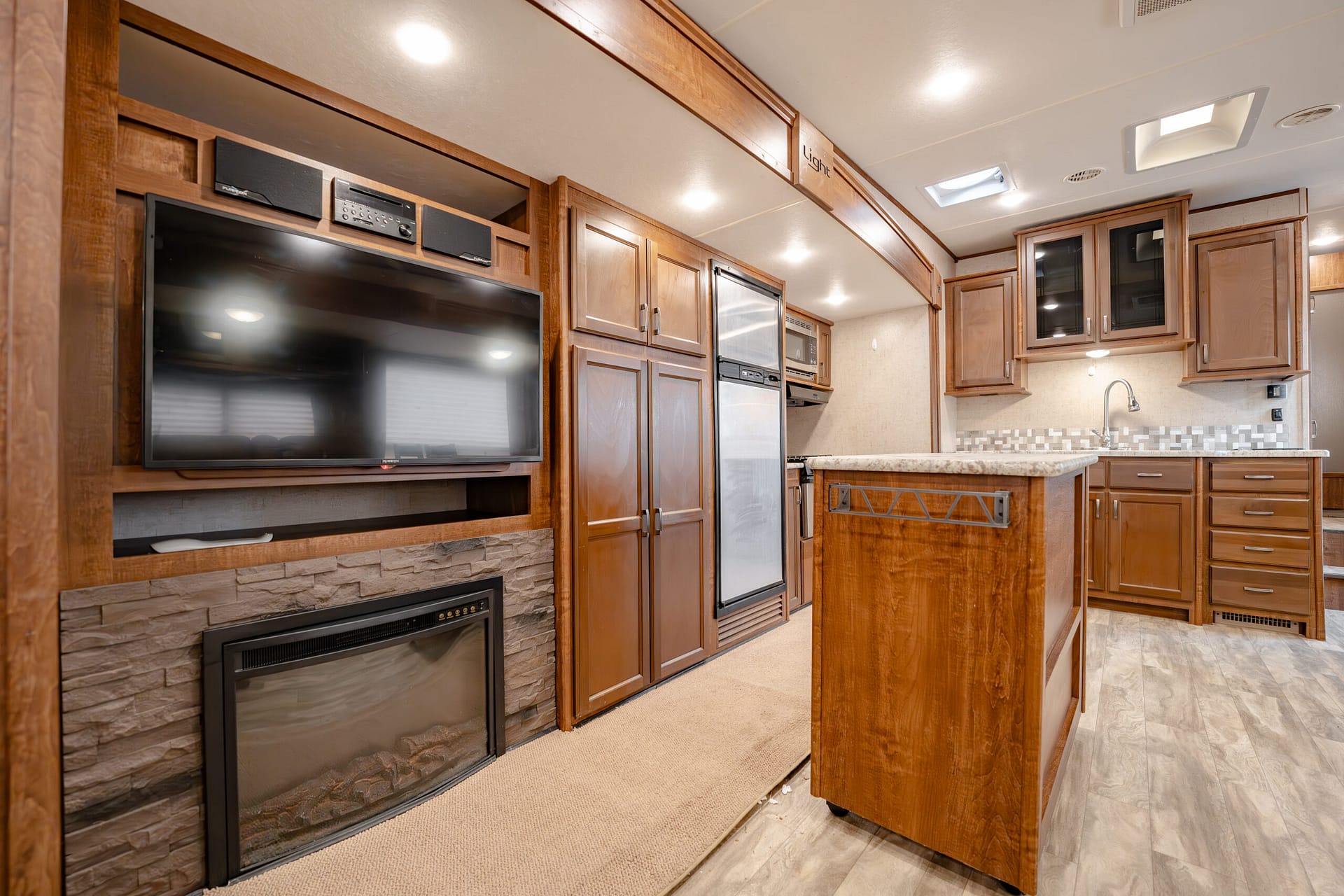 RV kitchen with warm wood cabinets, island, stainless steel fridge, and a wall-mounted TV above a stone fireplace.