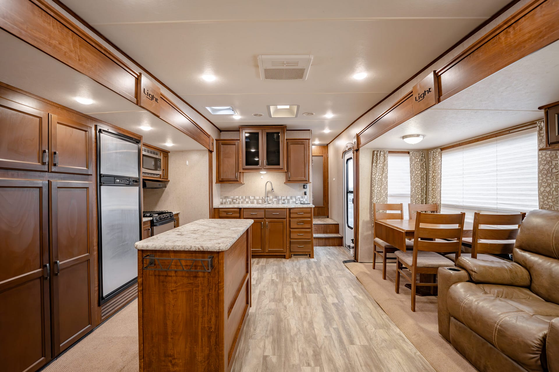 Interior of a modern RV: wood cabinets, stainless fridge, and a central kitchen island next to a dining area.