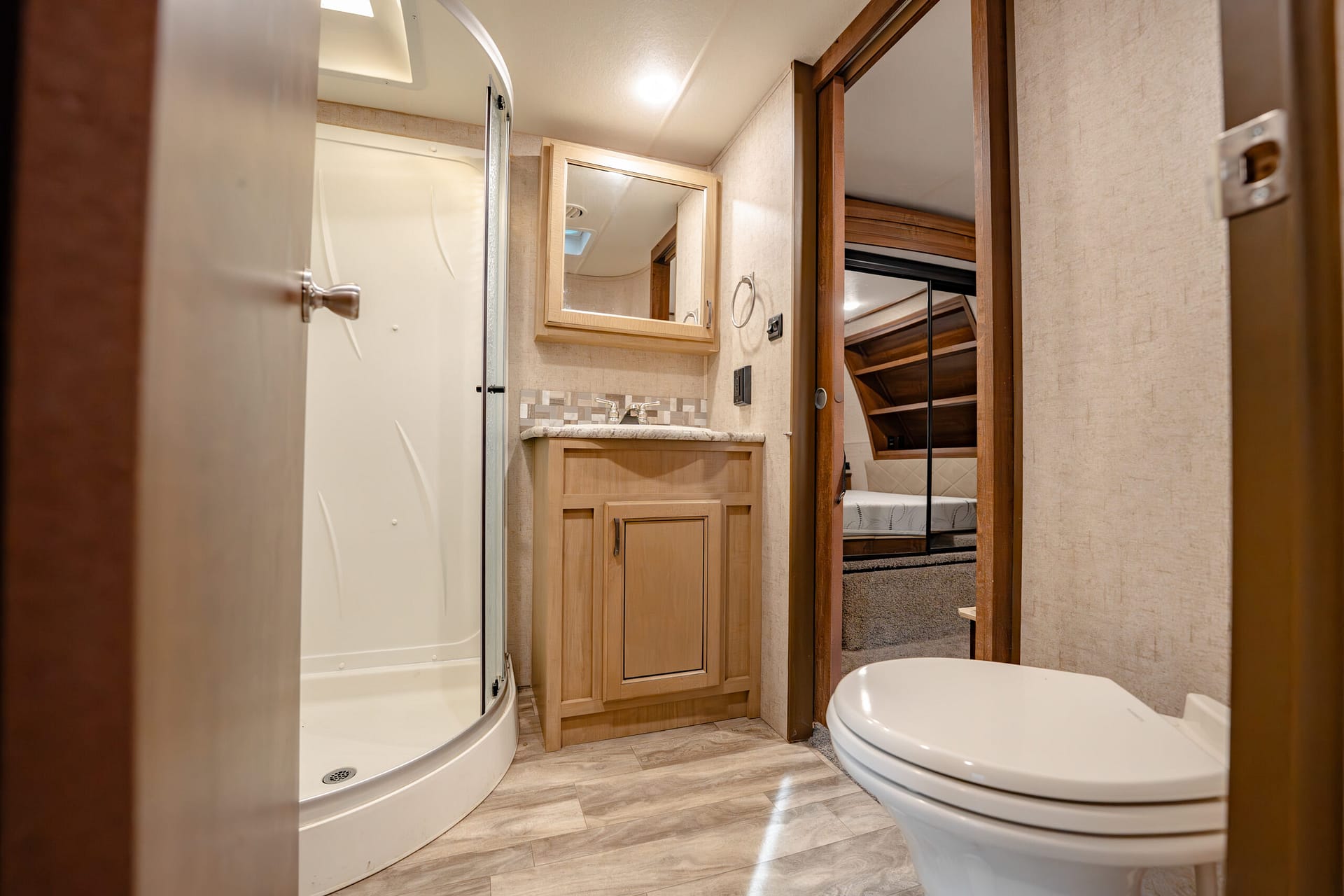 RV bathroom with curved corner shower, wooden vanity, and mirror reflecting ceiling light. A toilet sits to the right and a doorway leads to a bedroom area.