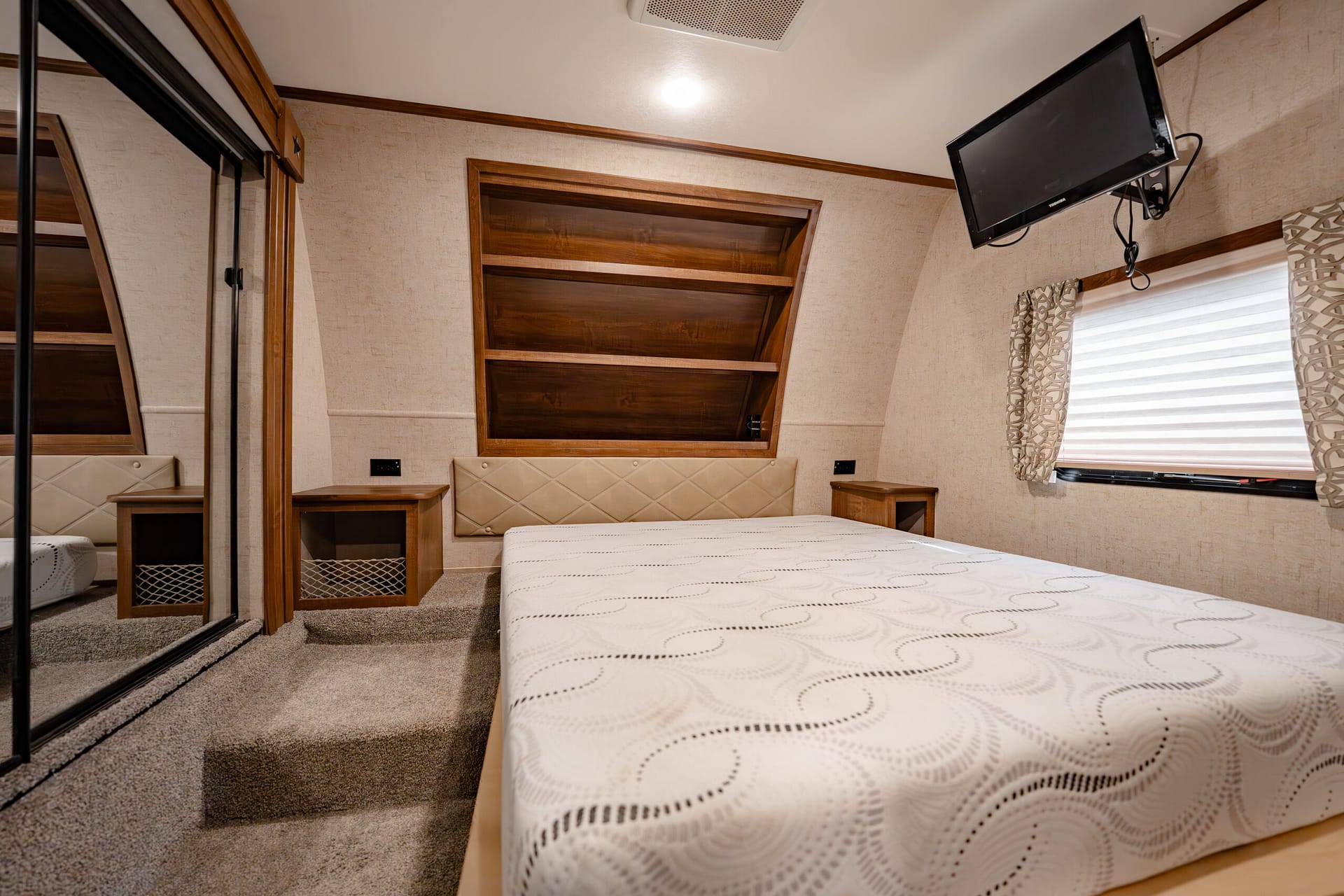 Bedroom inside a compact RV or trailer: center bed with quilted headboard, two small nightstands, built-in wooden shelves above, window with blinds, and a wall-mounted TV in the corner
