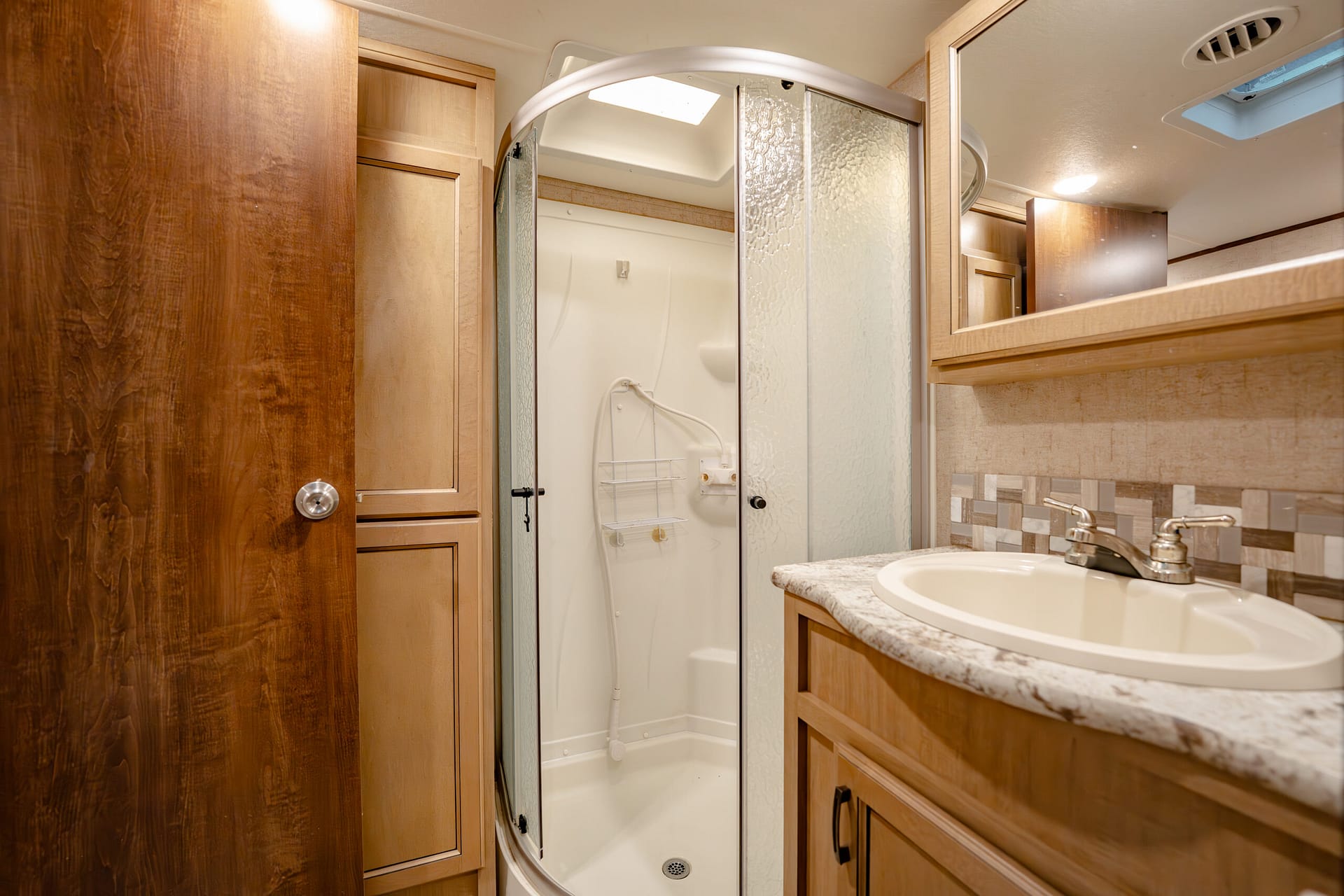 A compact bathroom with a curved frosted-glass shower enclosure, wooden cabinetry, and a marble countertop sink.