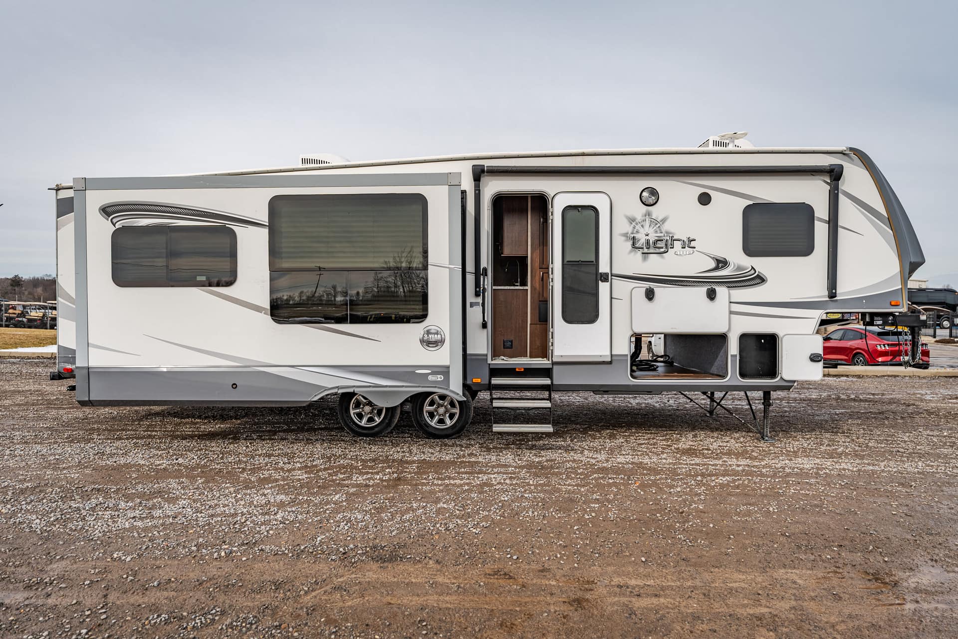 Modern white fifth-wheel travel trailer with a side slide-out, parked on a gravel lot; entry door is open with steps deployed and a visible interior doorway.