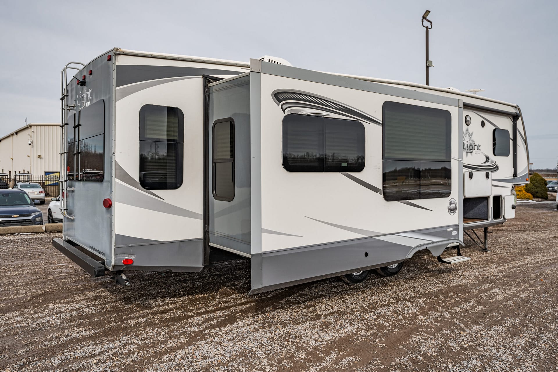 Travel trailer with an extended slide-out parked on a gravel lot, outdoors in front of a building and cars