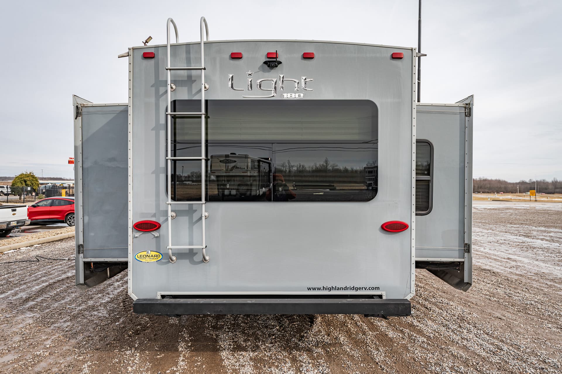 Rear view of a silver travel trailer with a metal ladder and large rear window, parked on a gravel lot.