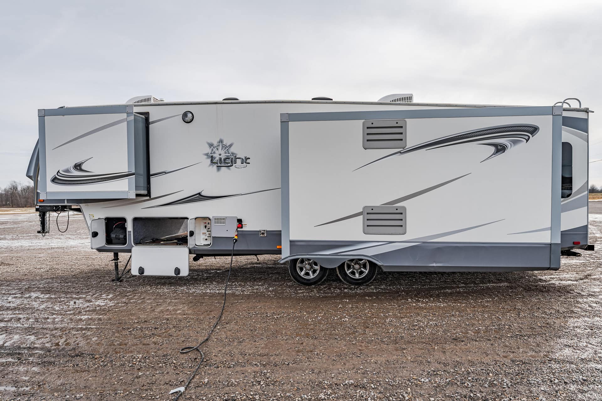 White travel trailer with a rear extended slide-out on a gravel lot; power cord plugged into the exterior outlet at the front left of the trailer.
