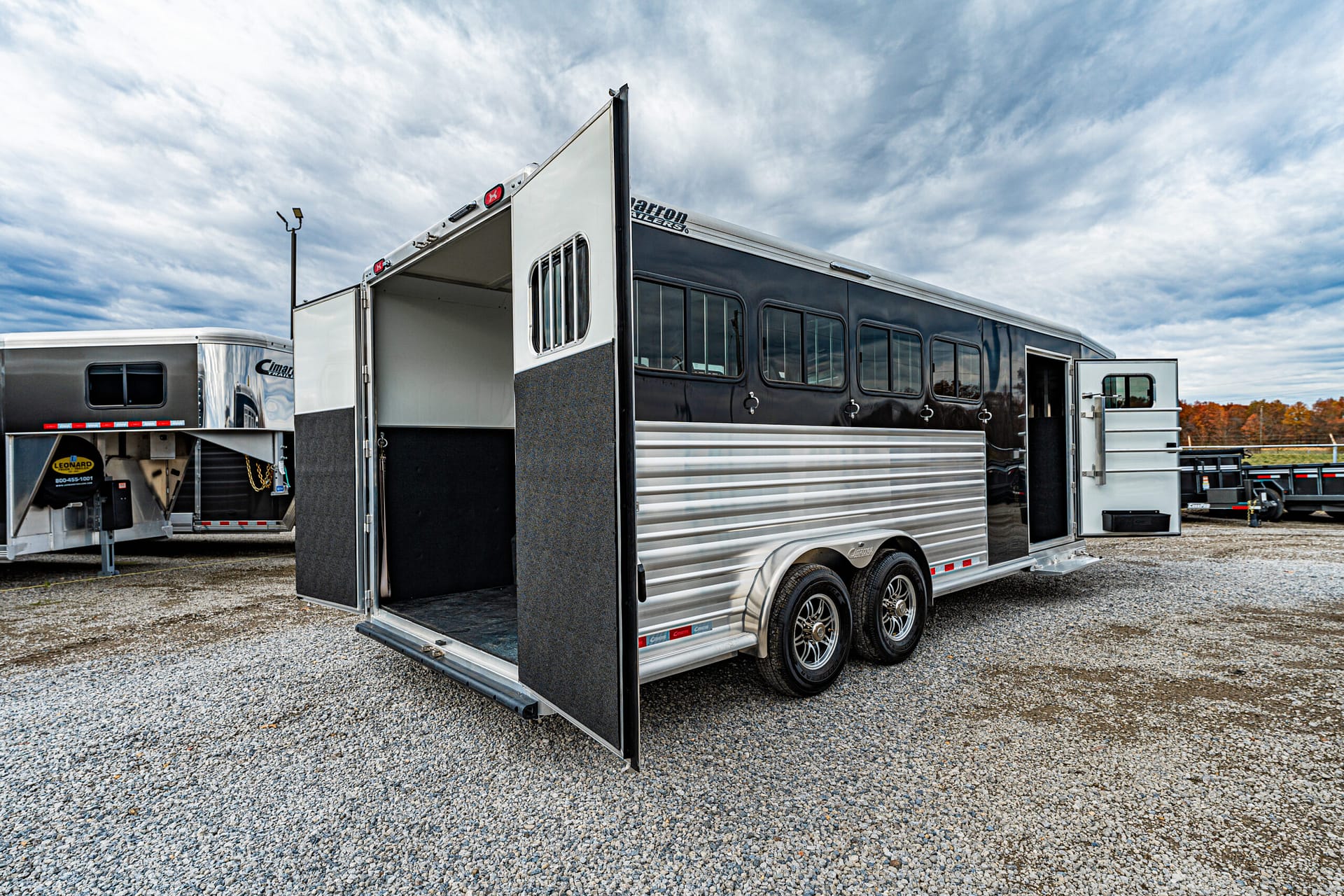Silver and black two-axle horse trailer with the rear ramp down, parked on a gravel lot with other trailers in the background under a cloudy sky.