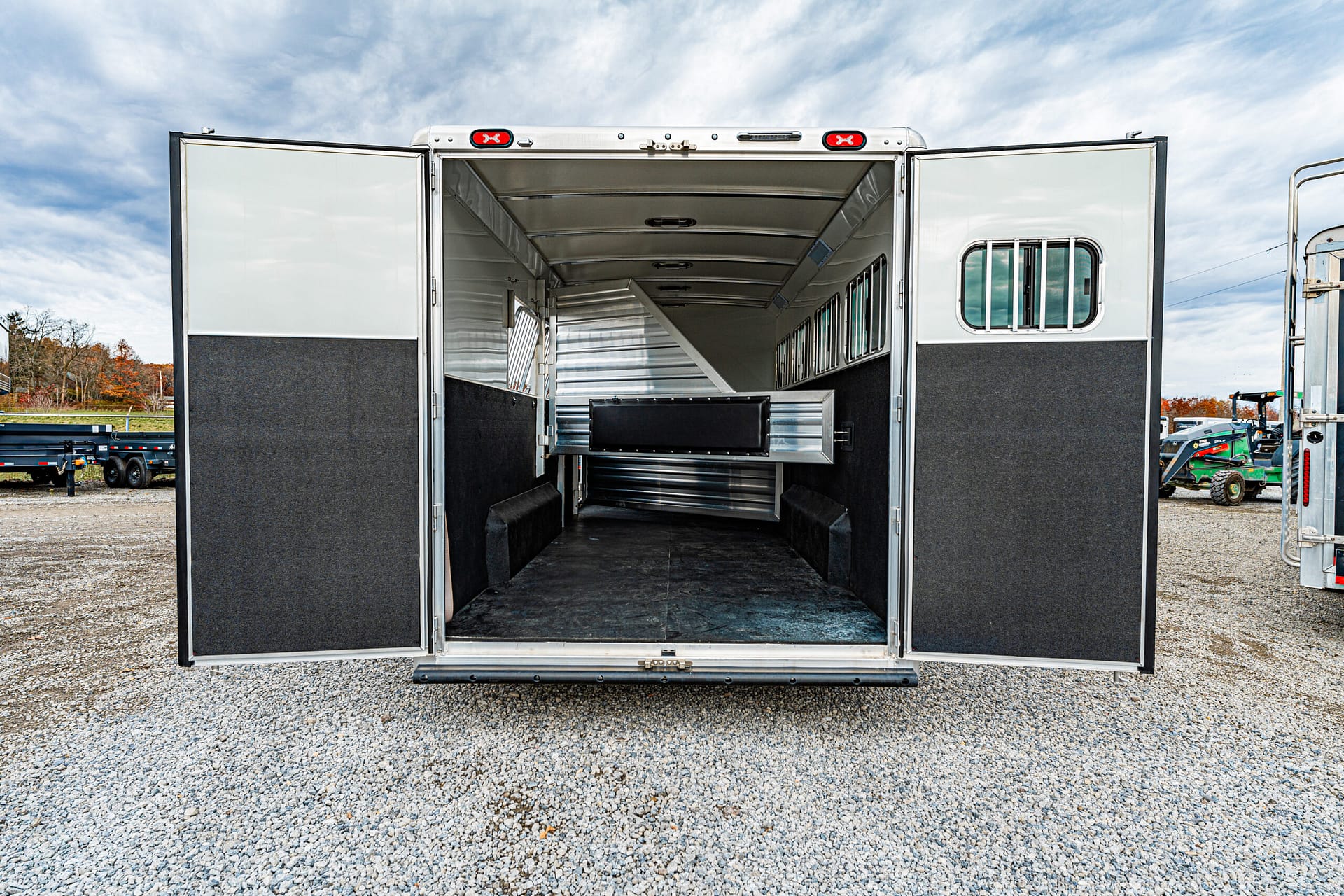 Rear view of an open horse trailer showing a central divider, padded walls, and a gravel lot outside.