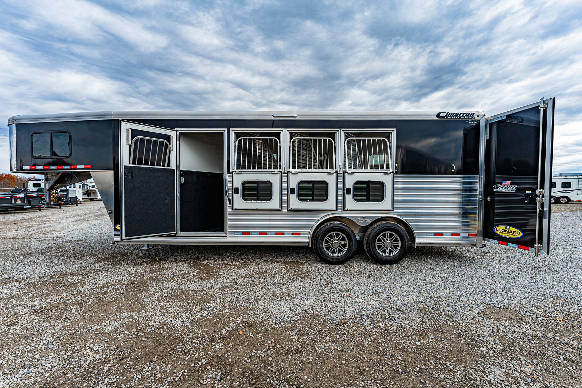Long aluminum horse trailer with three open stalls and a side entry, parked on gravel. Generated for transport of horses on a sunny day.