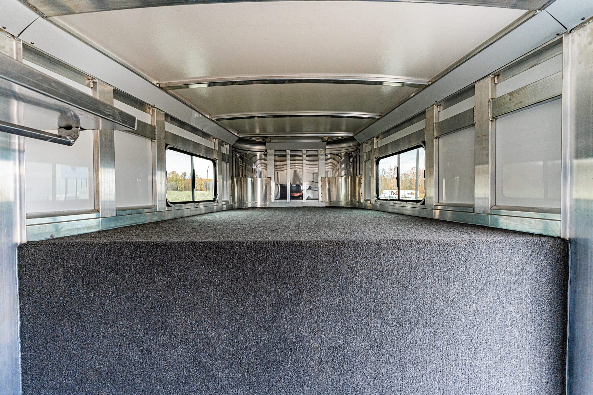 Interior of an empty aluminum trailer with a gray carpeted floor and side windows, viewed from the rear toward the front.