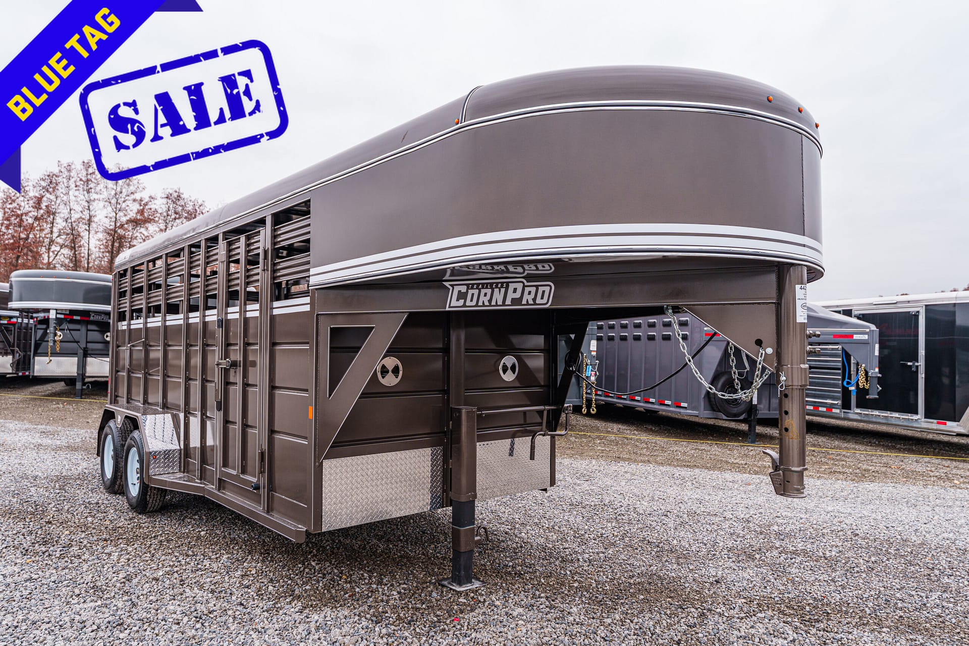 Brown gooseneck livestock trailer on a gravel lot with a blue 'Blue Tag Sale' banner overhead. The trailer has 'CornPro' branding on its side.