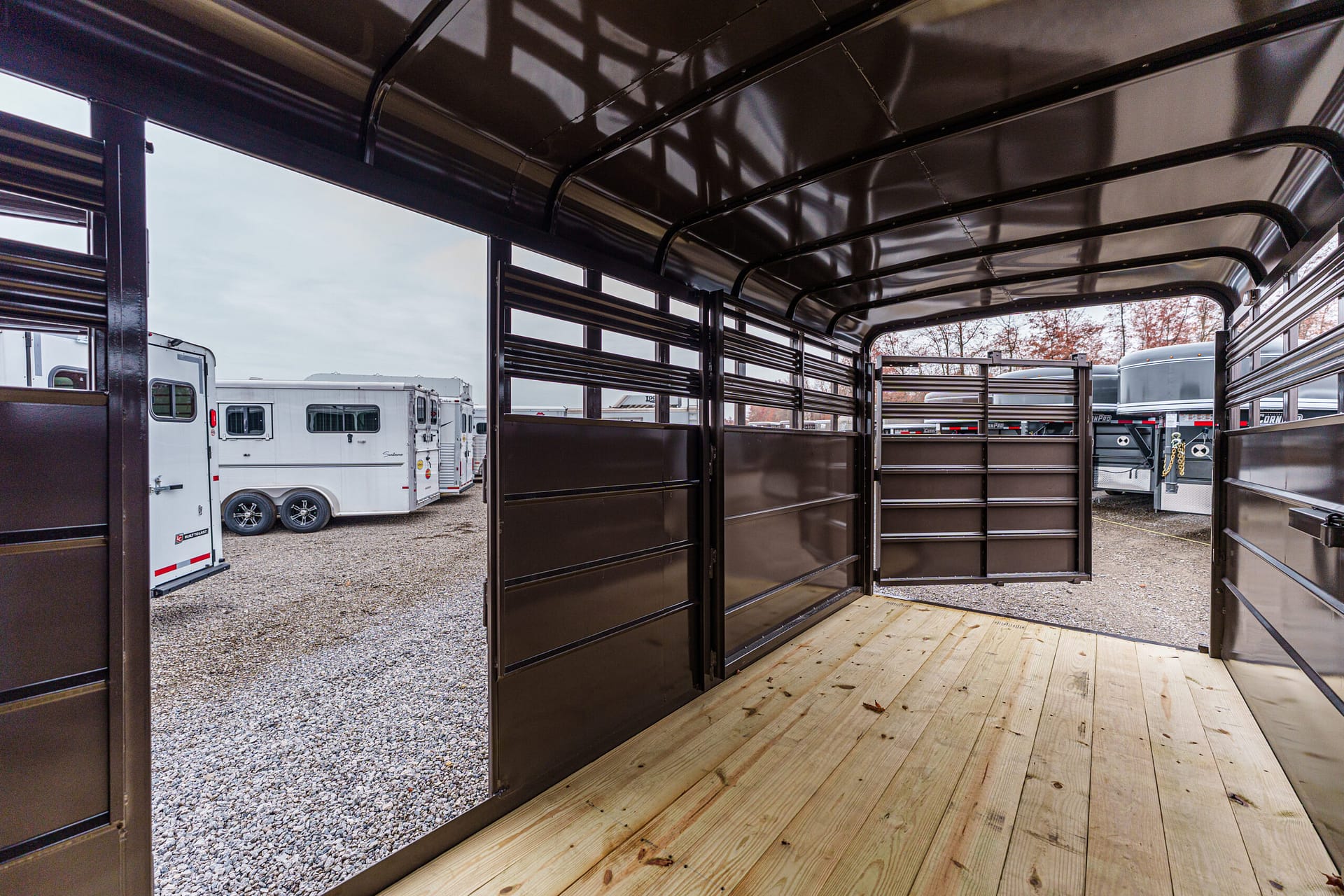 Interior of a black horse trailer with open side panels and wooden floor, other trailers visible outside on a gravel lot